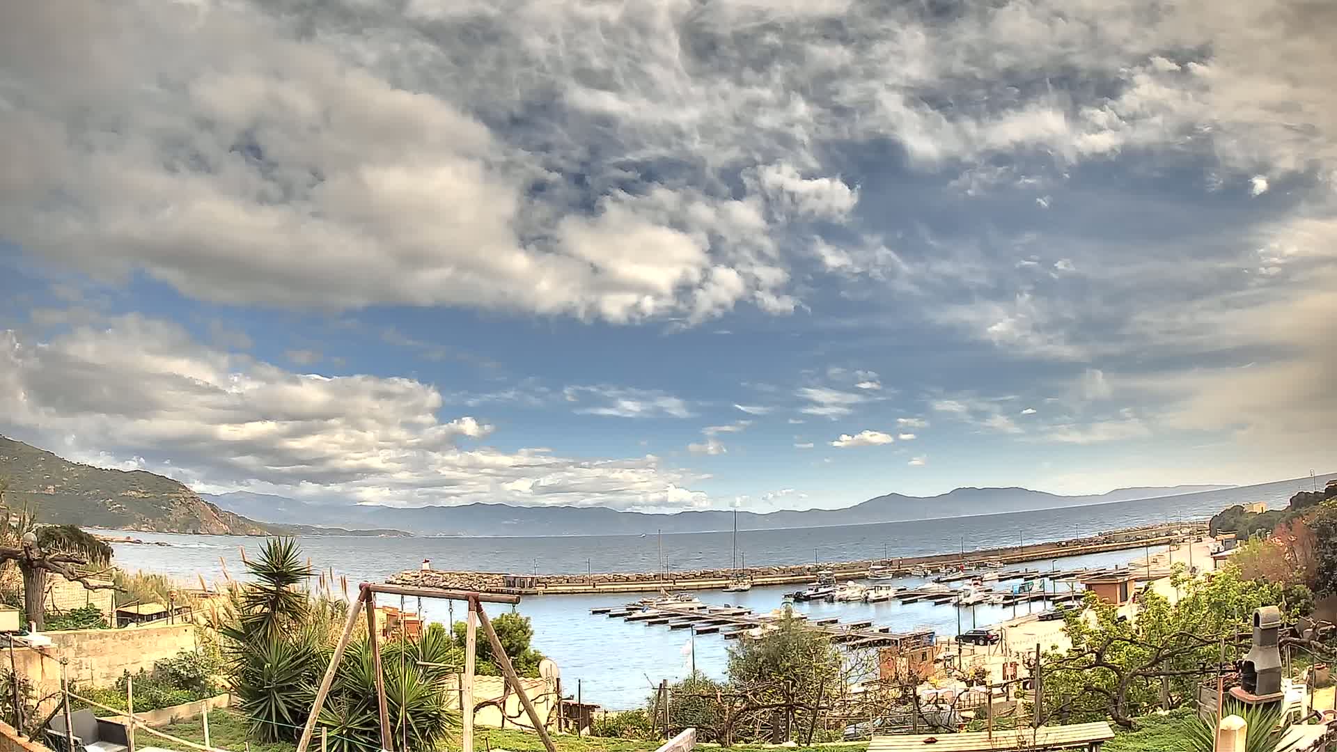 A partly cloudy day overlooking a calm bay filled with docked boats, mountains in the distance, and a small garden in the foreground.