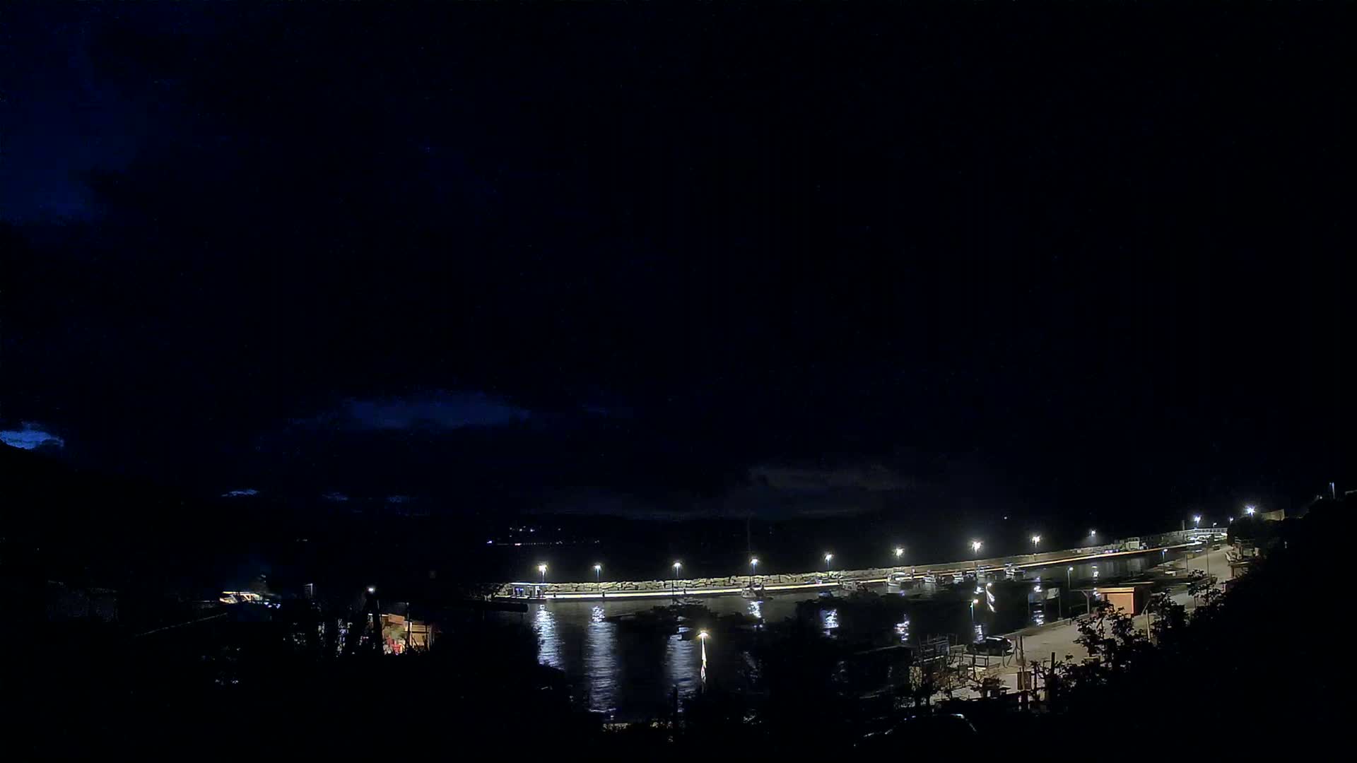 A nighttime view of a harbor with boats and lit docks under a dark, cloudy sky.