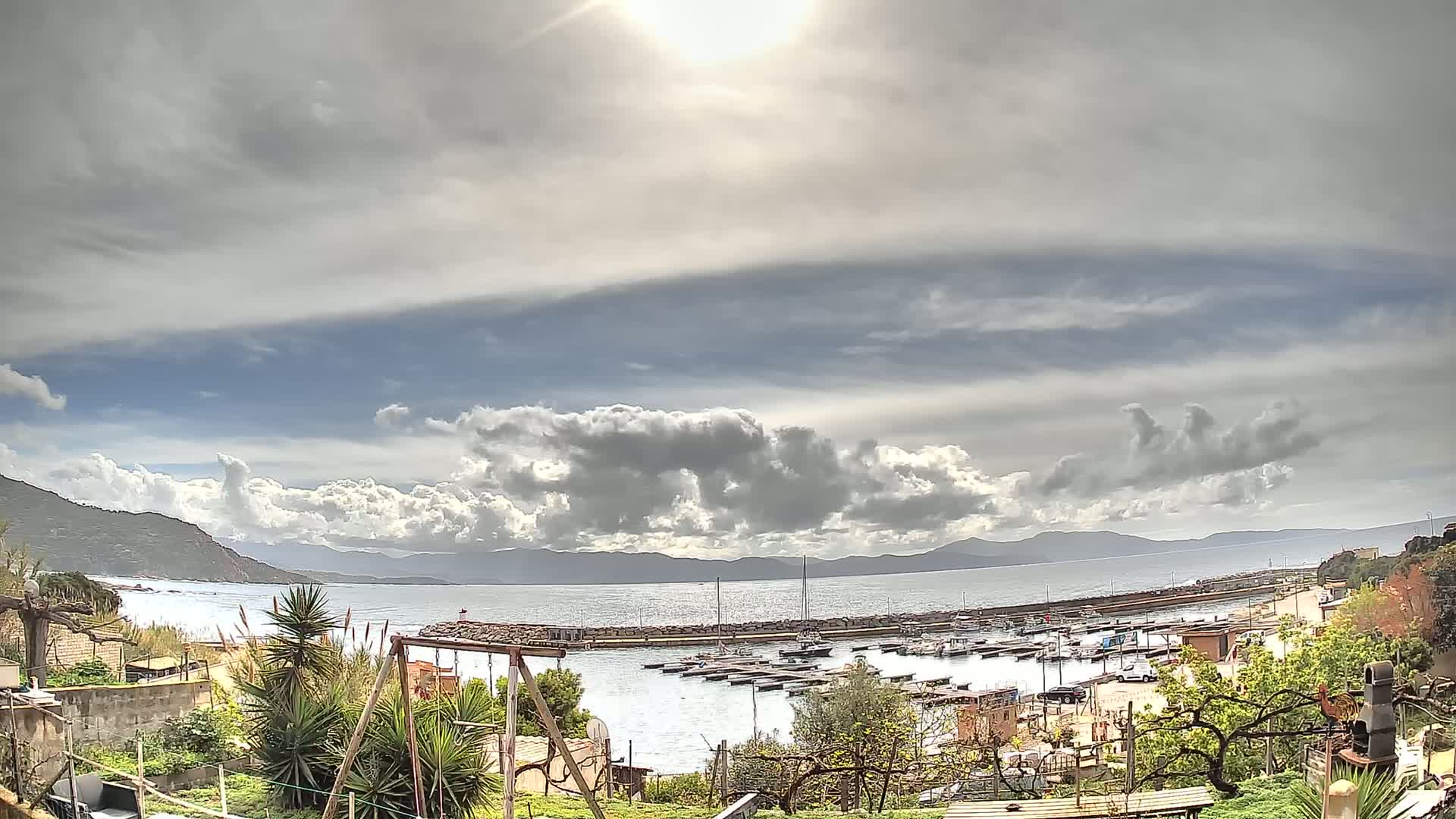 A partly sunny day with puffy clouds over a calm bay filled with boats, mountains in the distance, and a small garden in the foreground.