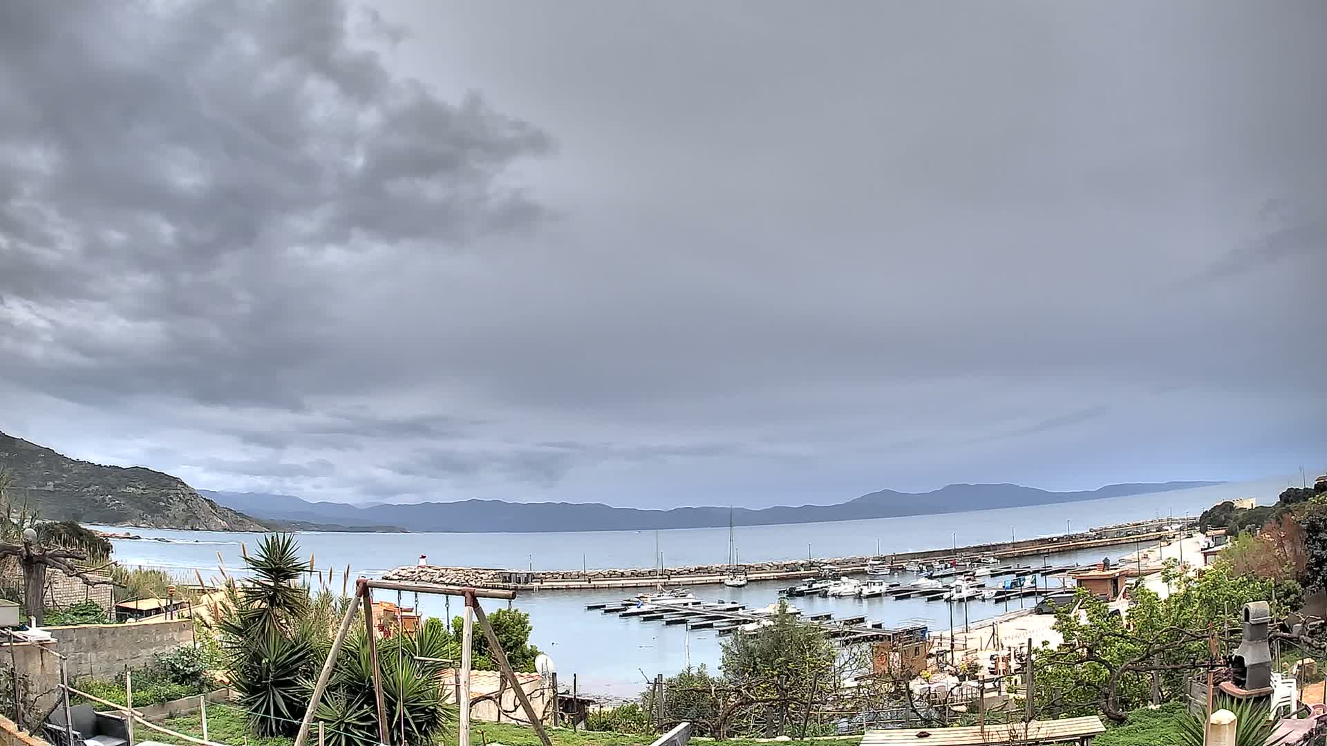 A small harbor filled with boats is seen from a hilltop overlooking a calm sea under a cloudy sky.