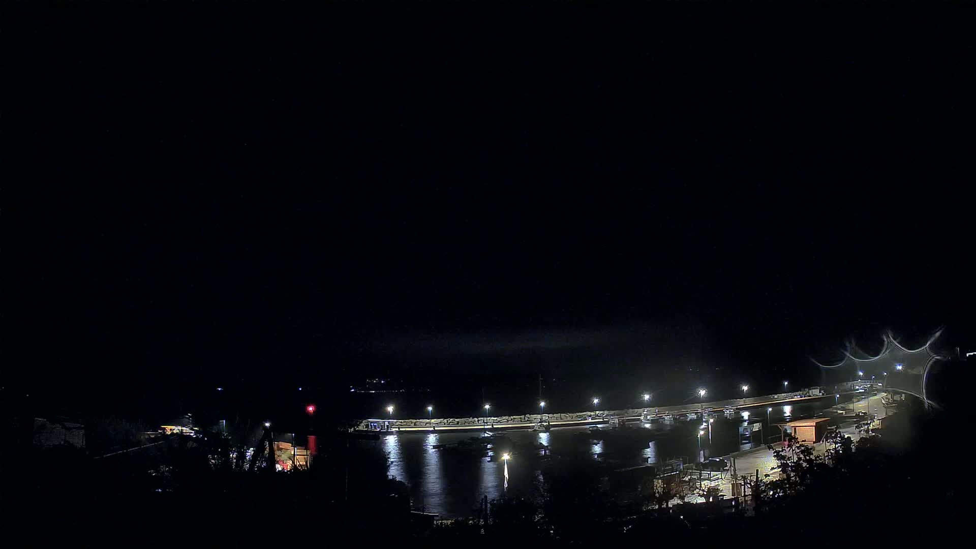 A nighttime view of a harbor with boats moored at a pier, illuminated by lights under a dark, clear sky.