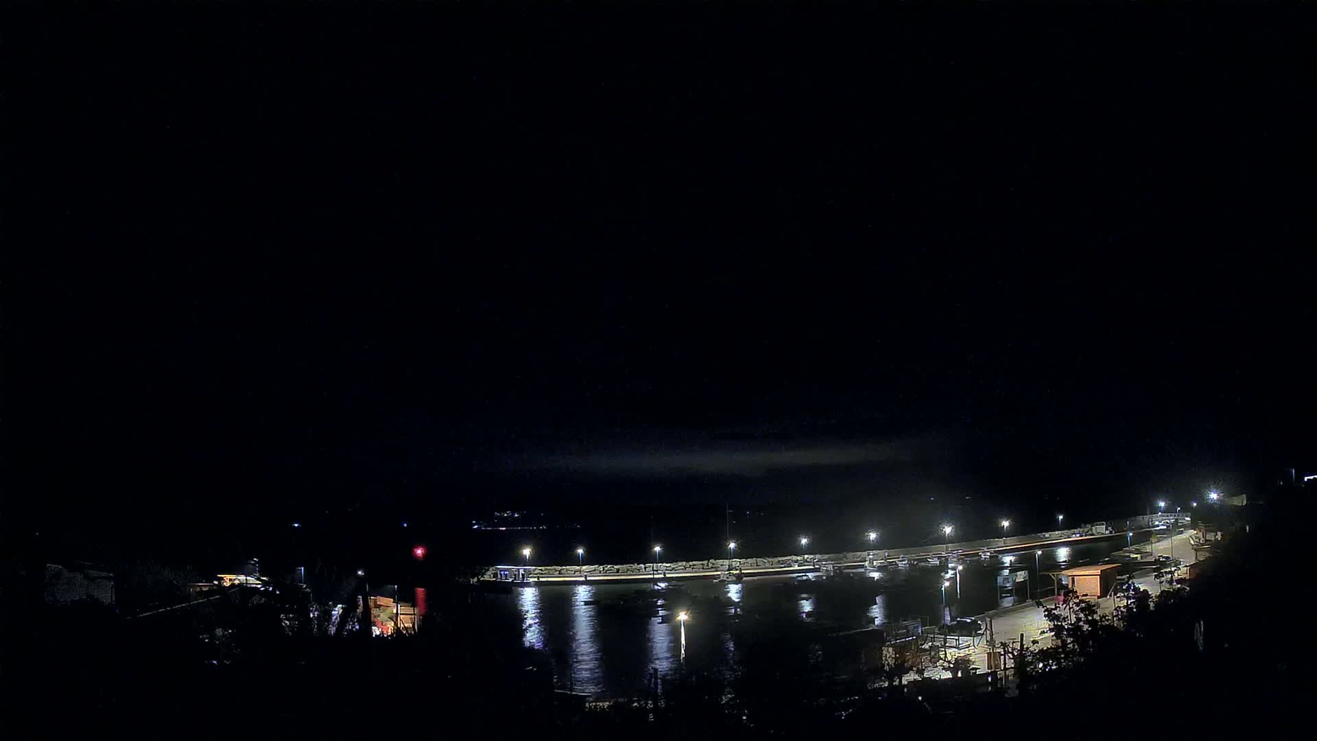 A nighttime view of a harbor with several boats moored, illuminated by lights along a seawall under a dark, cloudy sky.