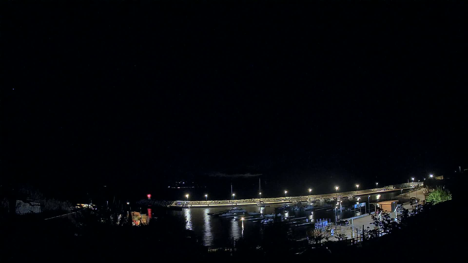 A nighttime view of a harbor filled with boats, illuminated by lights along a stone wall under a clear, dark sky.