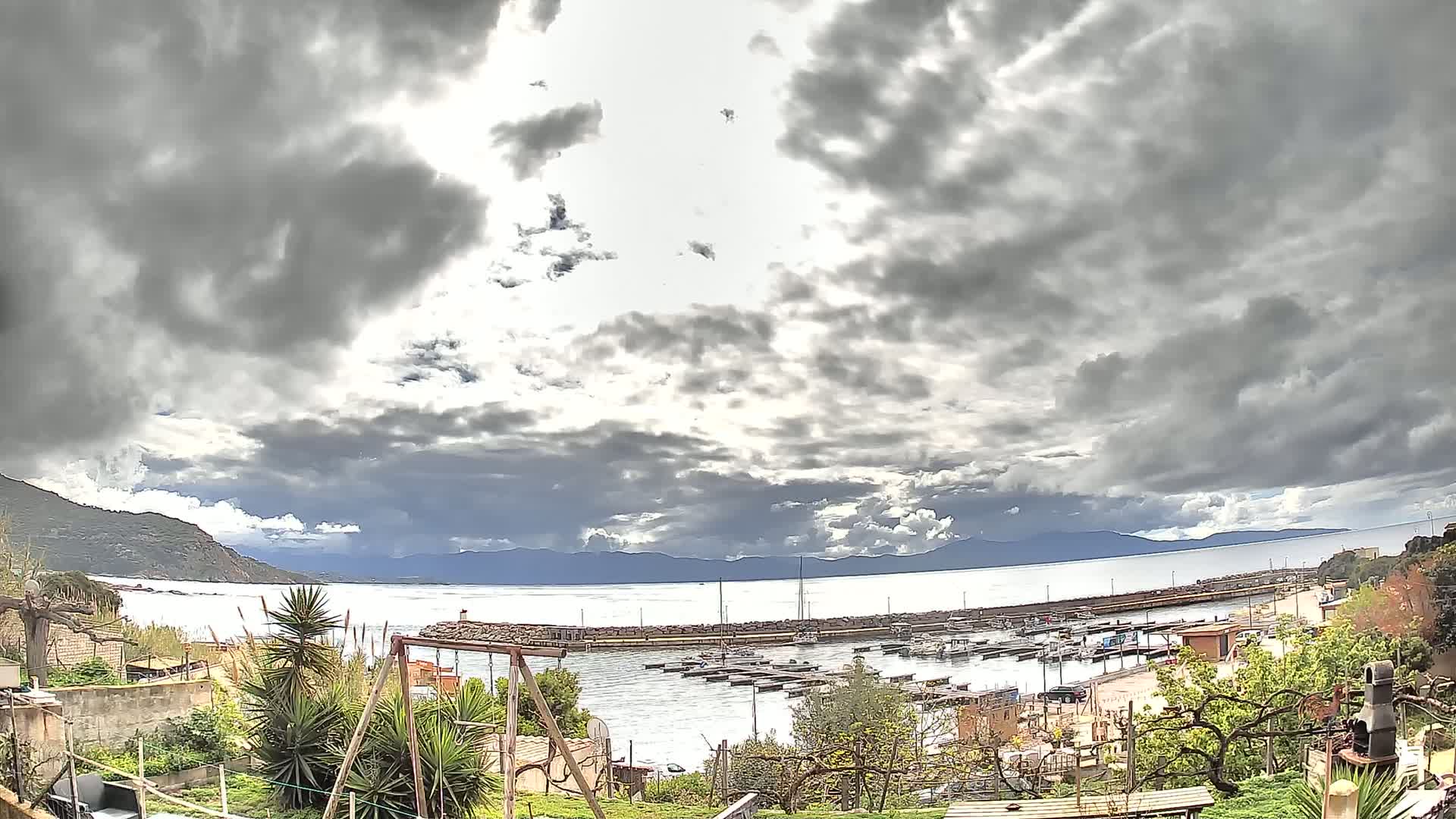 A small harbor filled with boats is seen from a hillside garden under a cloudy sky.