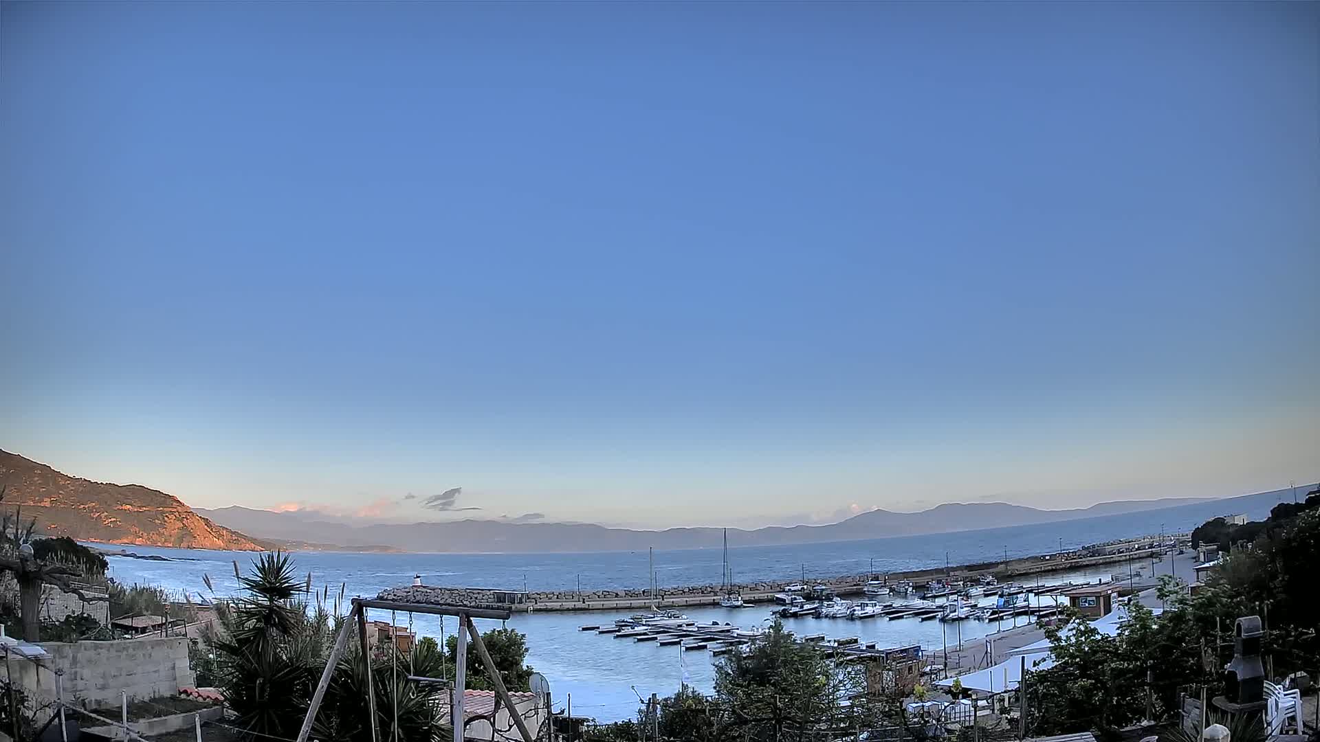 A small harbor filled with boats is nestled between a rocky coastline and calm ocean under a clear blue sky.
