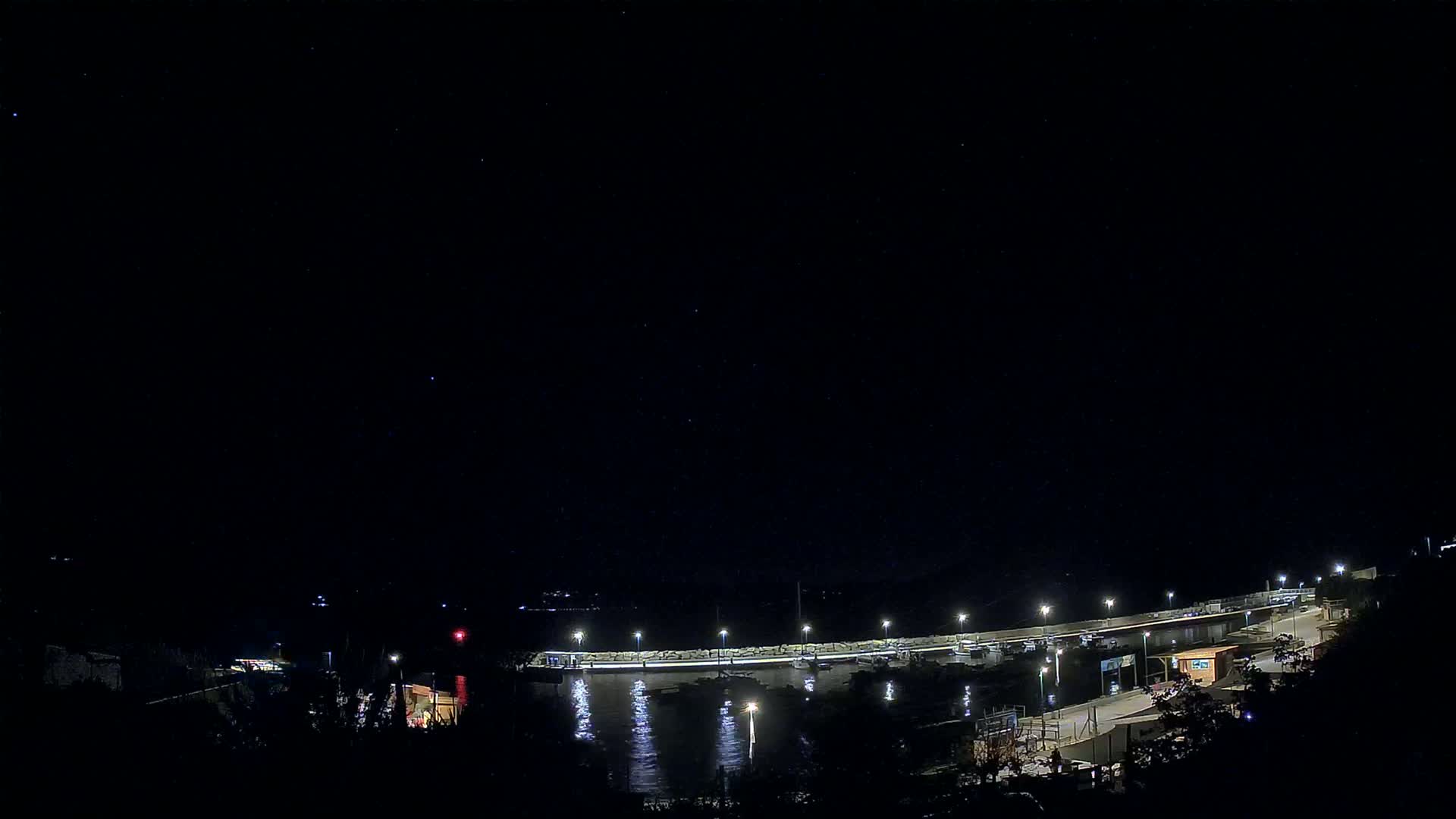 A nighttime view of a harbor with boats moored along a lit-up pier under a clear, starry sky.