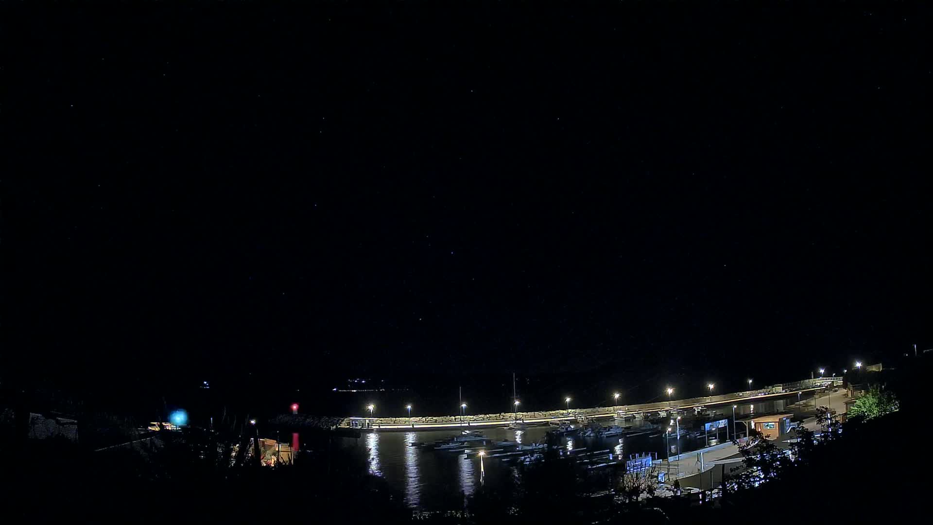 A nighttime view of a harbor with docked boats, lit by artificial lights under a clear, starry sky.