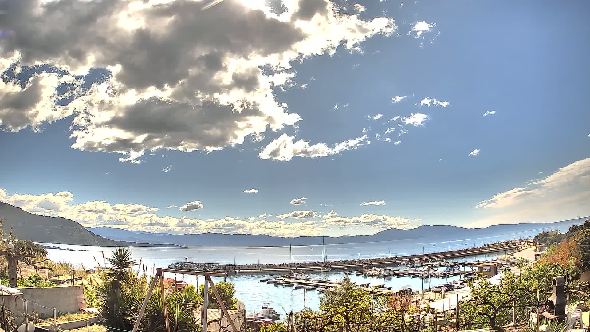 A partly sunny day shows a calm harbor filled with boats, backed by mountains under a partly cloudy sky.