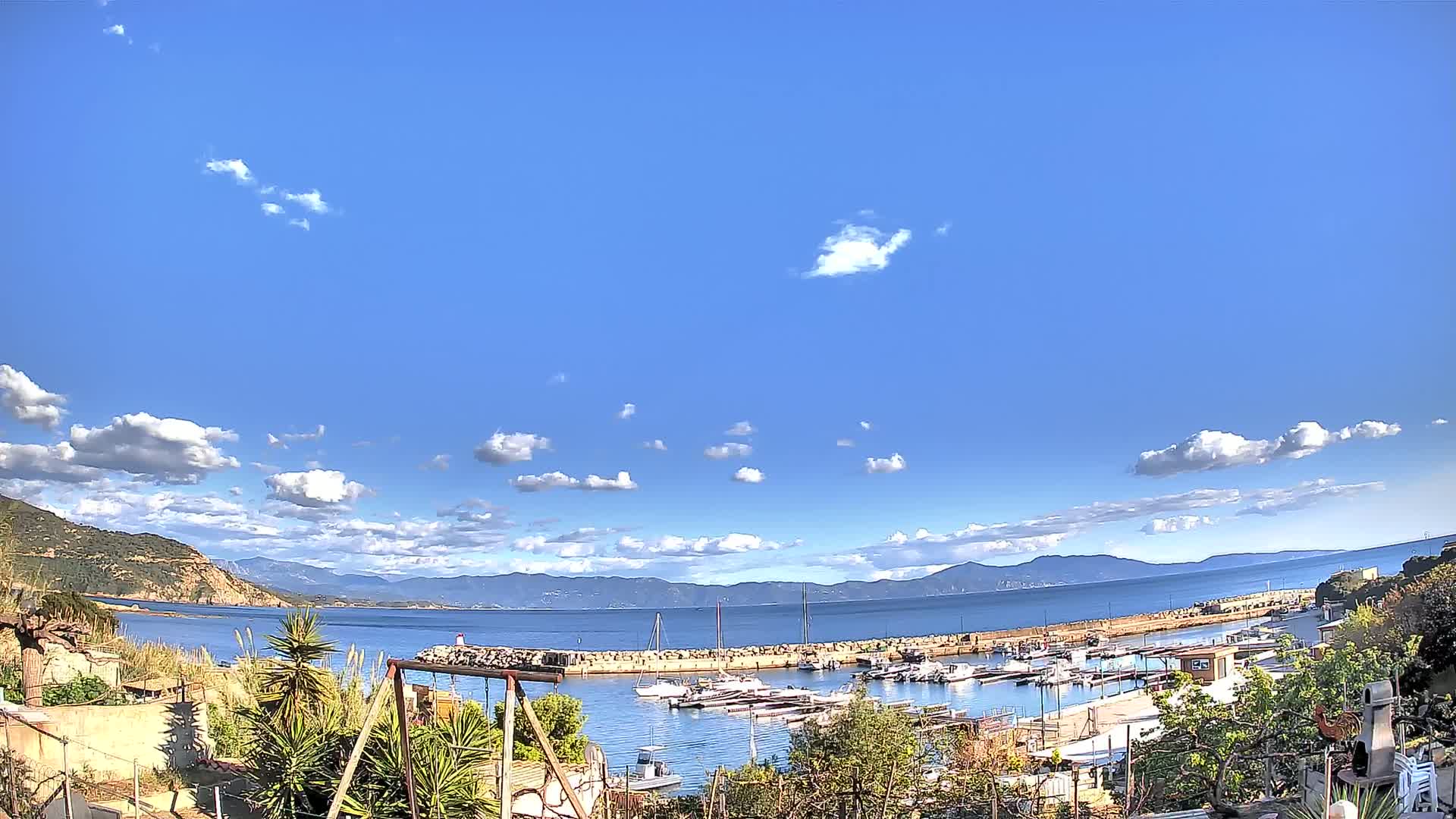 A small harbor filled with boats is visible under a mostly clear blue sky with scattered cumulus clouds, backed by a calm sea and distant mountains.