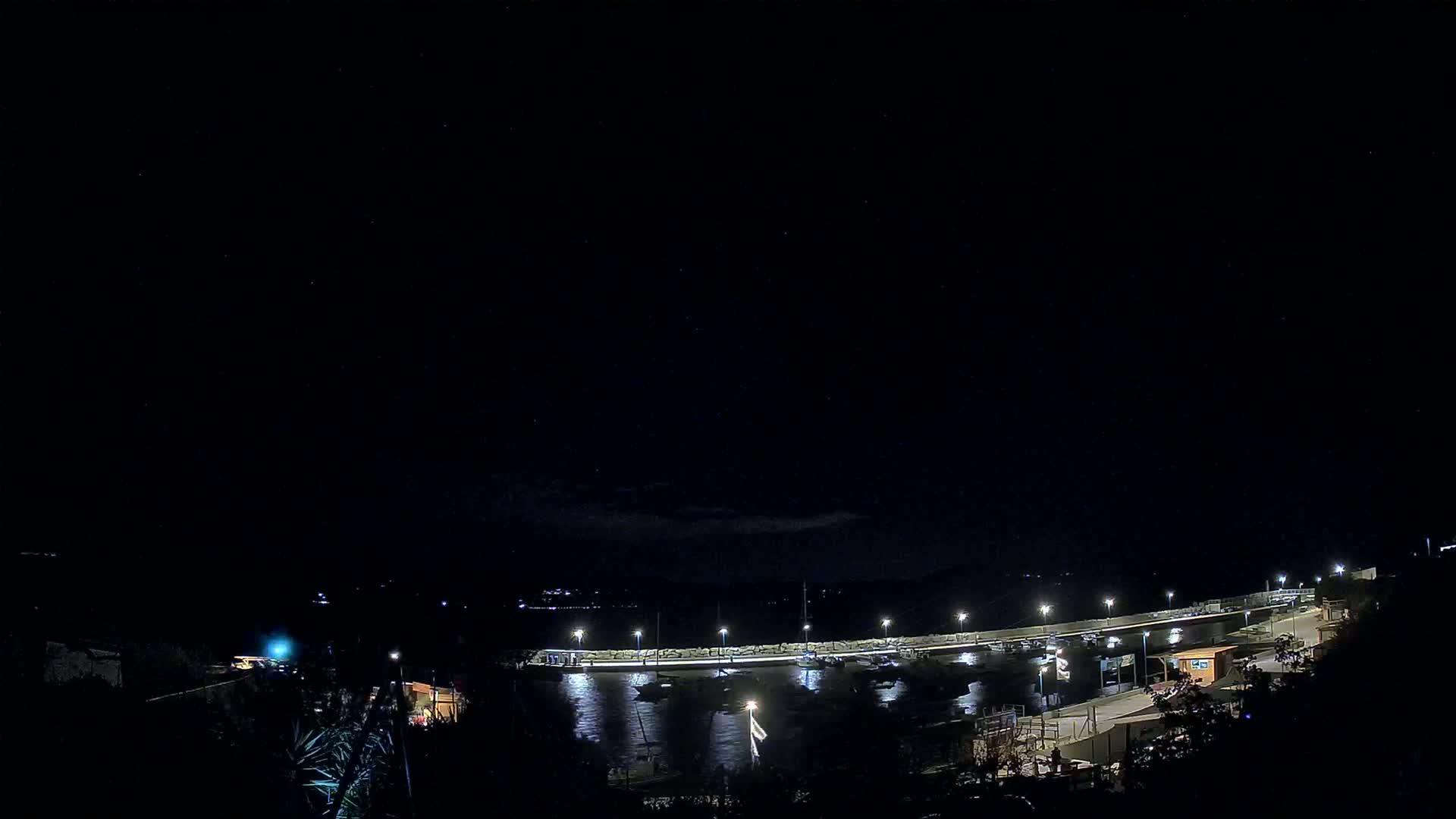 A nighttime view of a harbor with boats moored alongside a lit-up pier under a clear, starry sky.