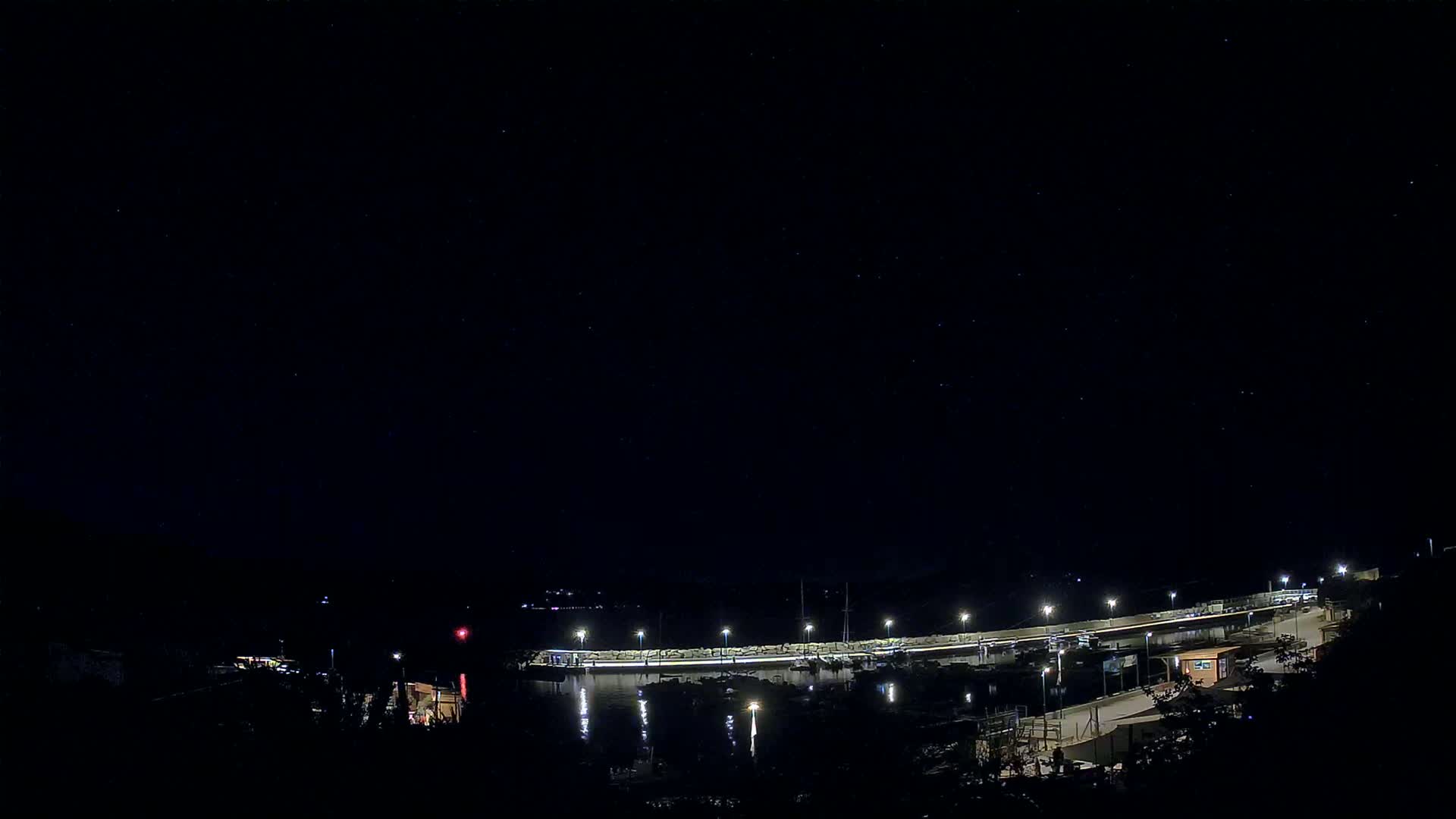 A nighttime view of a lit harbor with boats, a breakwater, and surrounding dark land under a clear, starry sky.