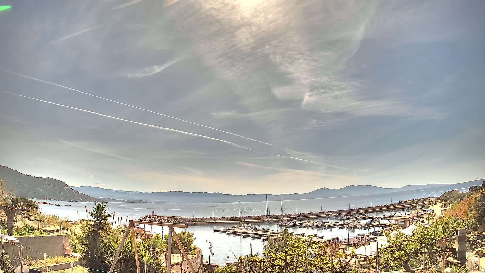 A partly cloudy day reveals a harbor filled with boats, nestled between hills and a calm sea under a bright sun.