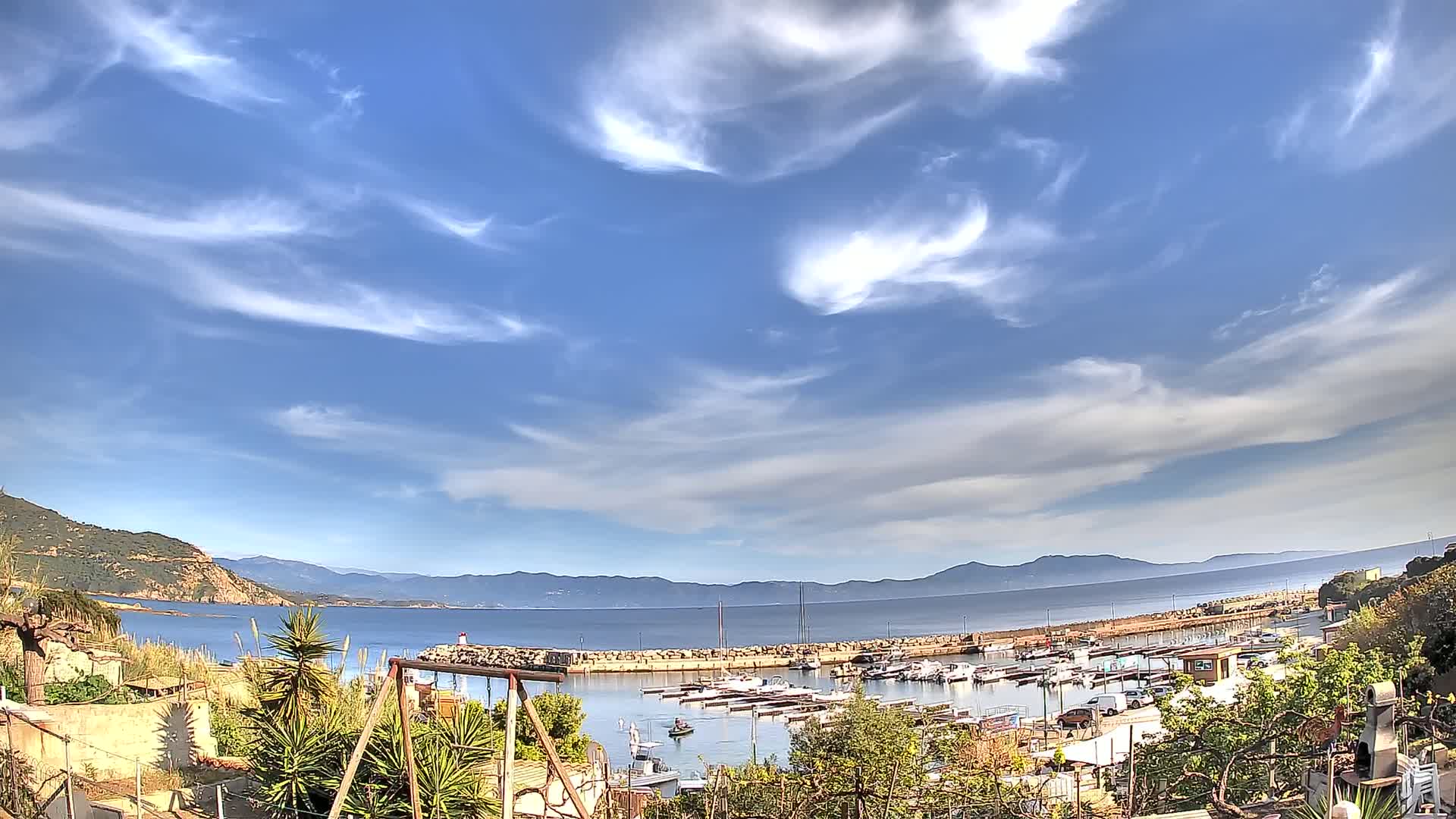 A harbor filled with boats is nestled between hills under a mostly sunny sky with wispy clouds.