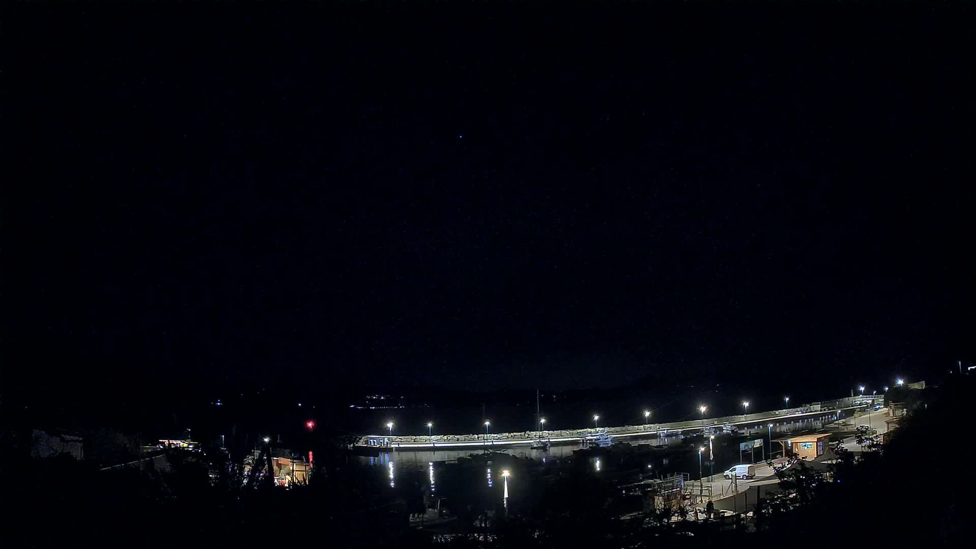 A nighttime, high-angle view shows a marina with boats and lit docks under a clear, dark sky.
