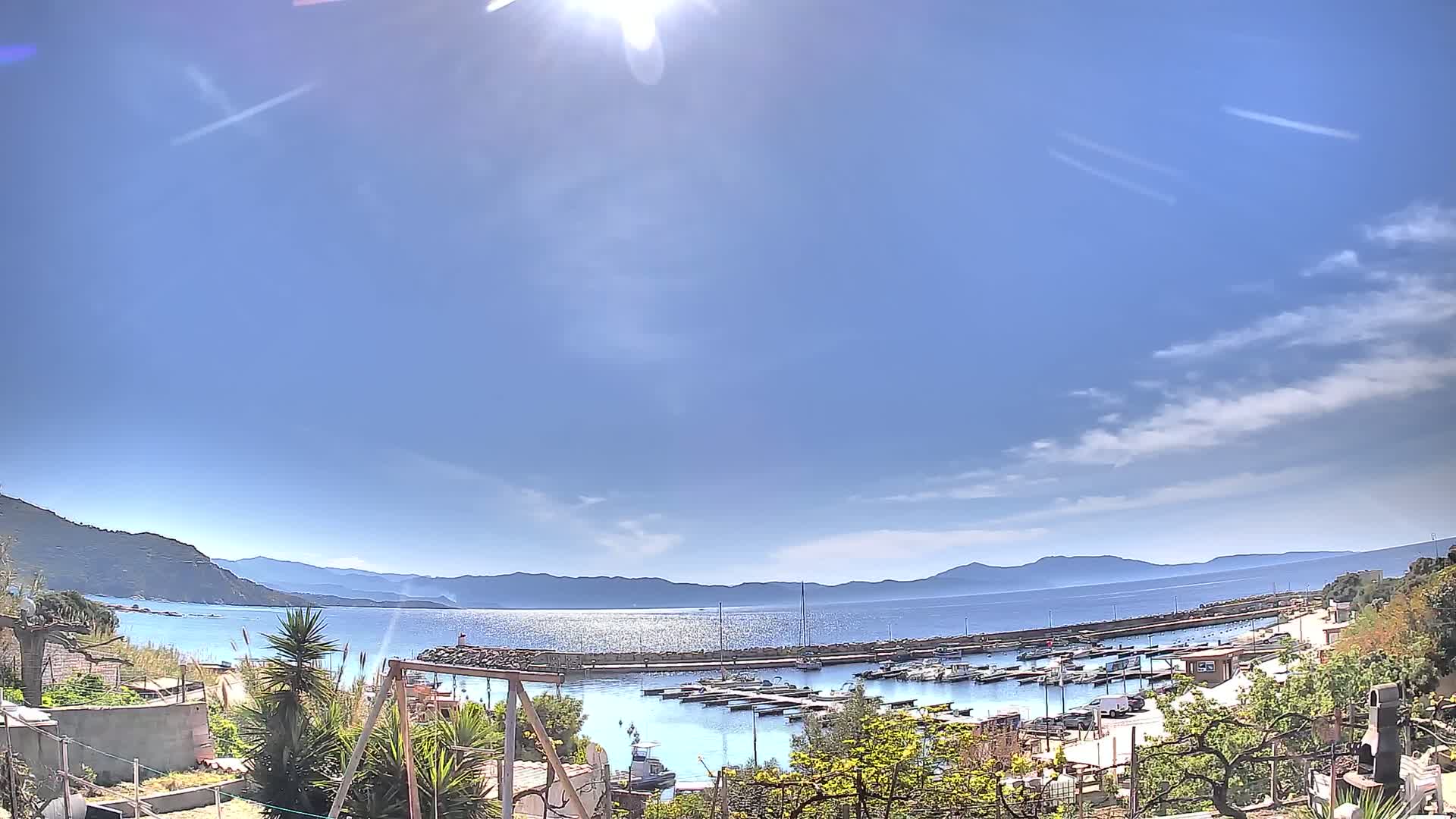 A sunny day reveals a calm harbor filled with boats, nestled between hills and a sparkling sea under a mostly clear sky.