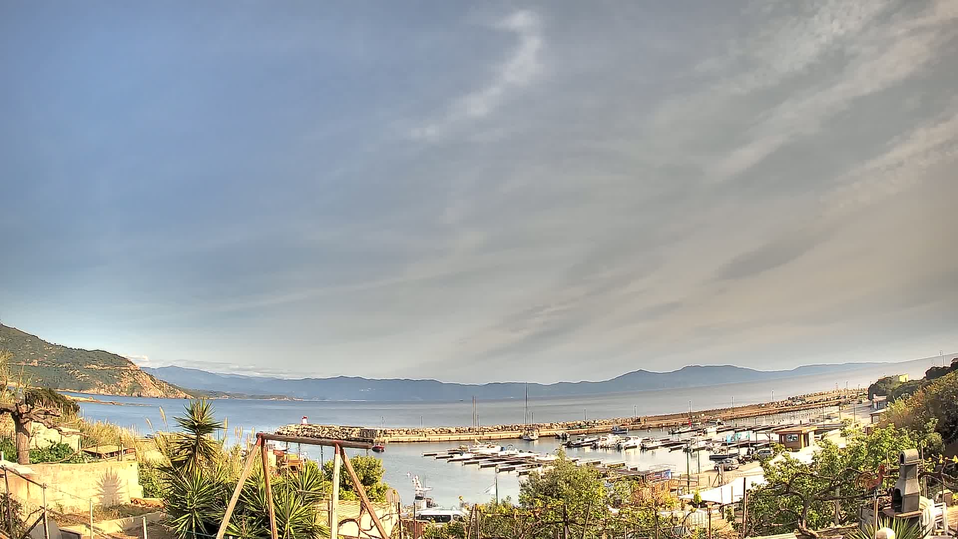 A small marina filled with boats is situated near a hillside, overlooking a calm bay and distant mountains under a partly cloudy sky.
