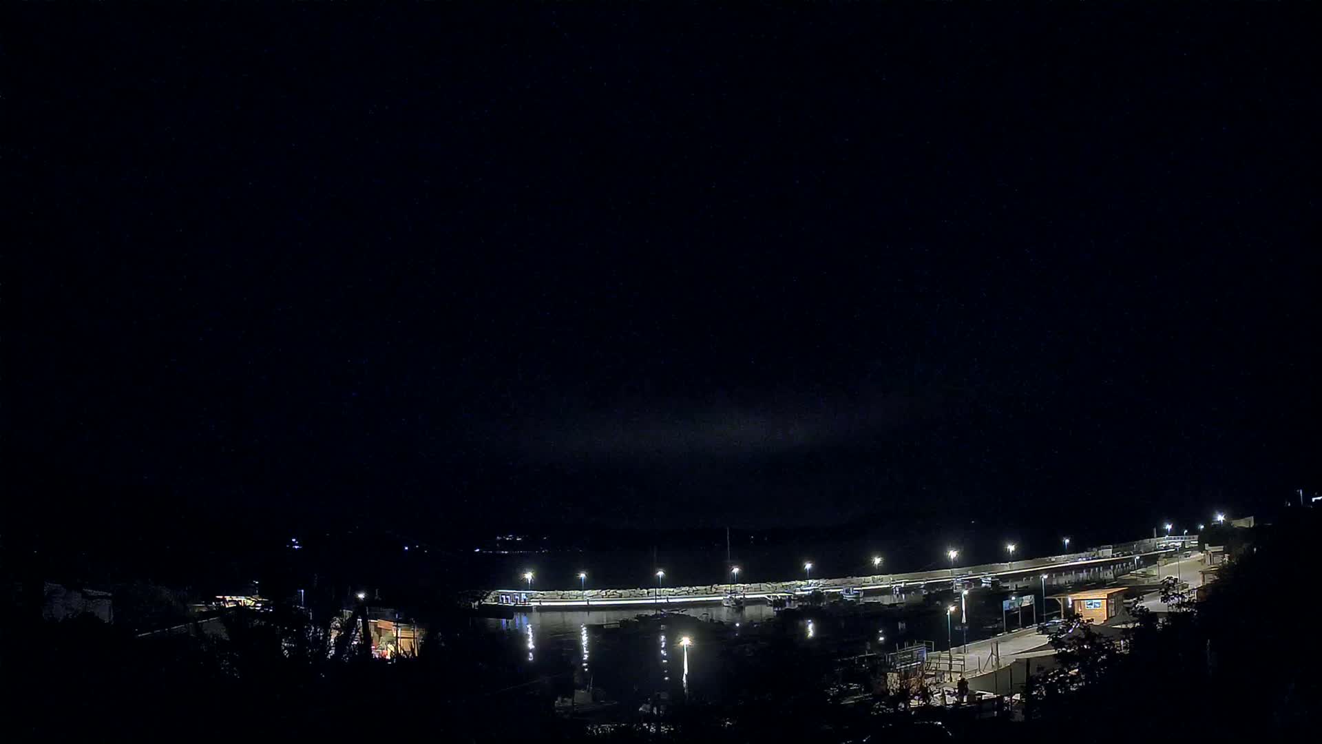 A nighttime view of a harbor with boats moored along a lit seawall under a dark, clear sky.