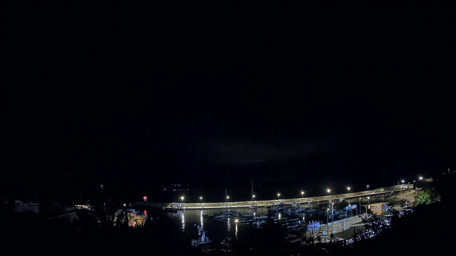 A nighttime view of a marina with numerous boats docked along a lit waterfront, under a dark, clear sky.