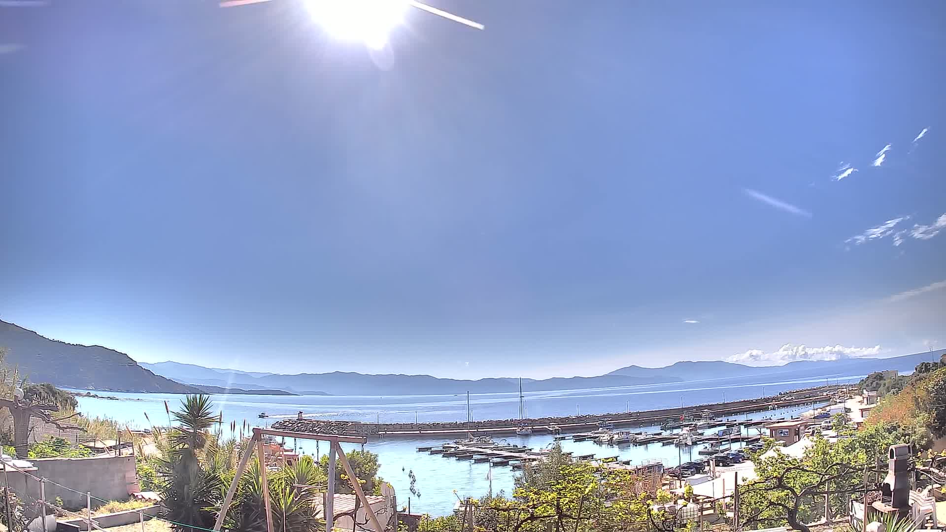 A sunny day overlooks a calm harbor filled with boats, nestled between hills and a clear blue sea under a mostly clear sky.