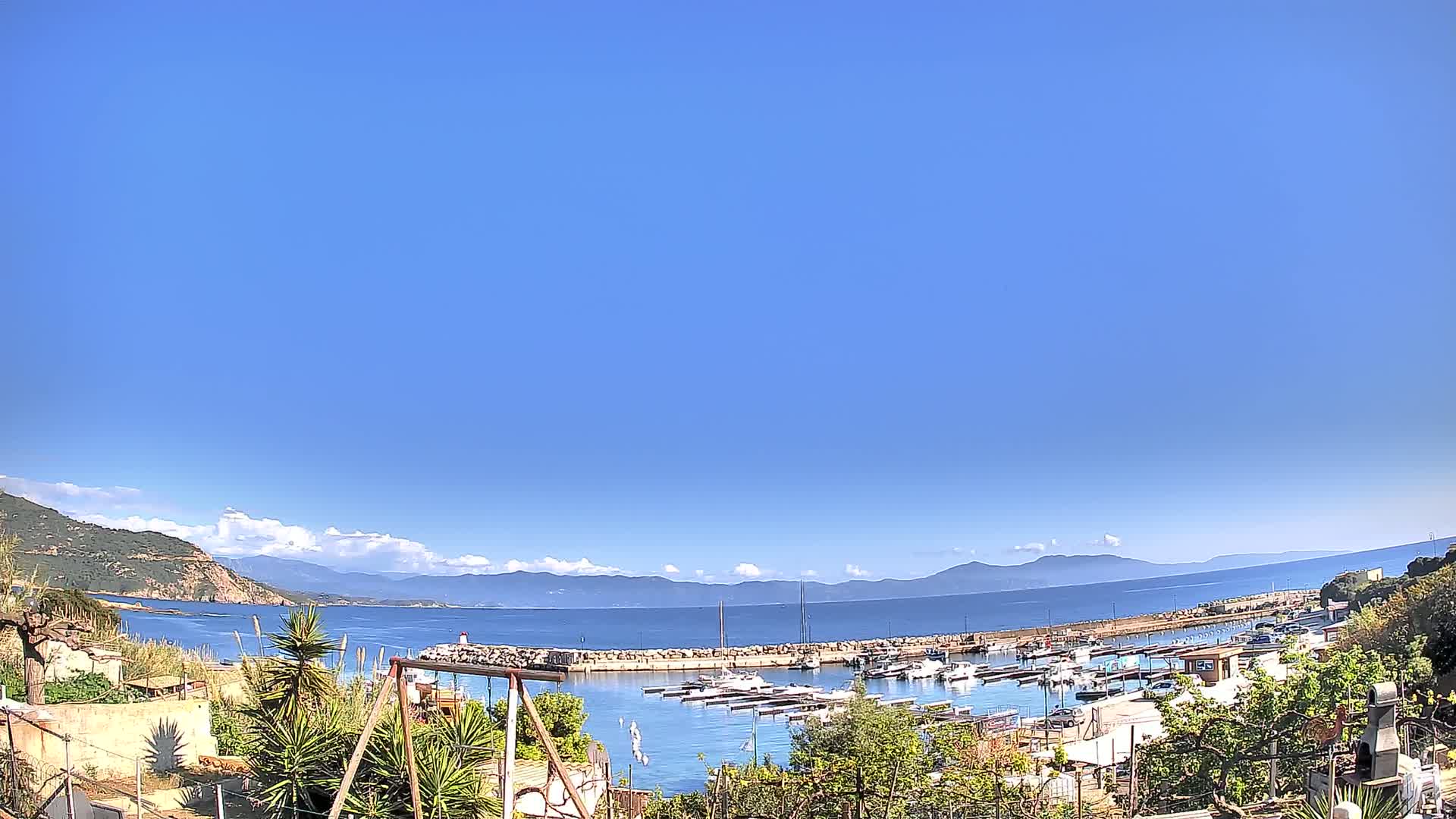 A small harbor filled with boats is nestled between a green hillside and a calm blue sea under a clear, bright sky.