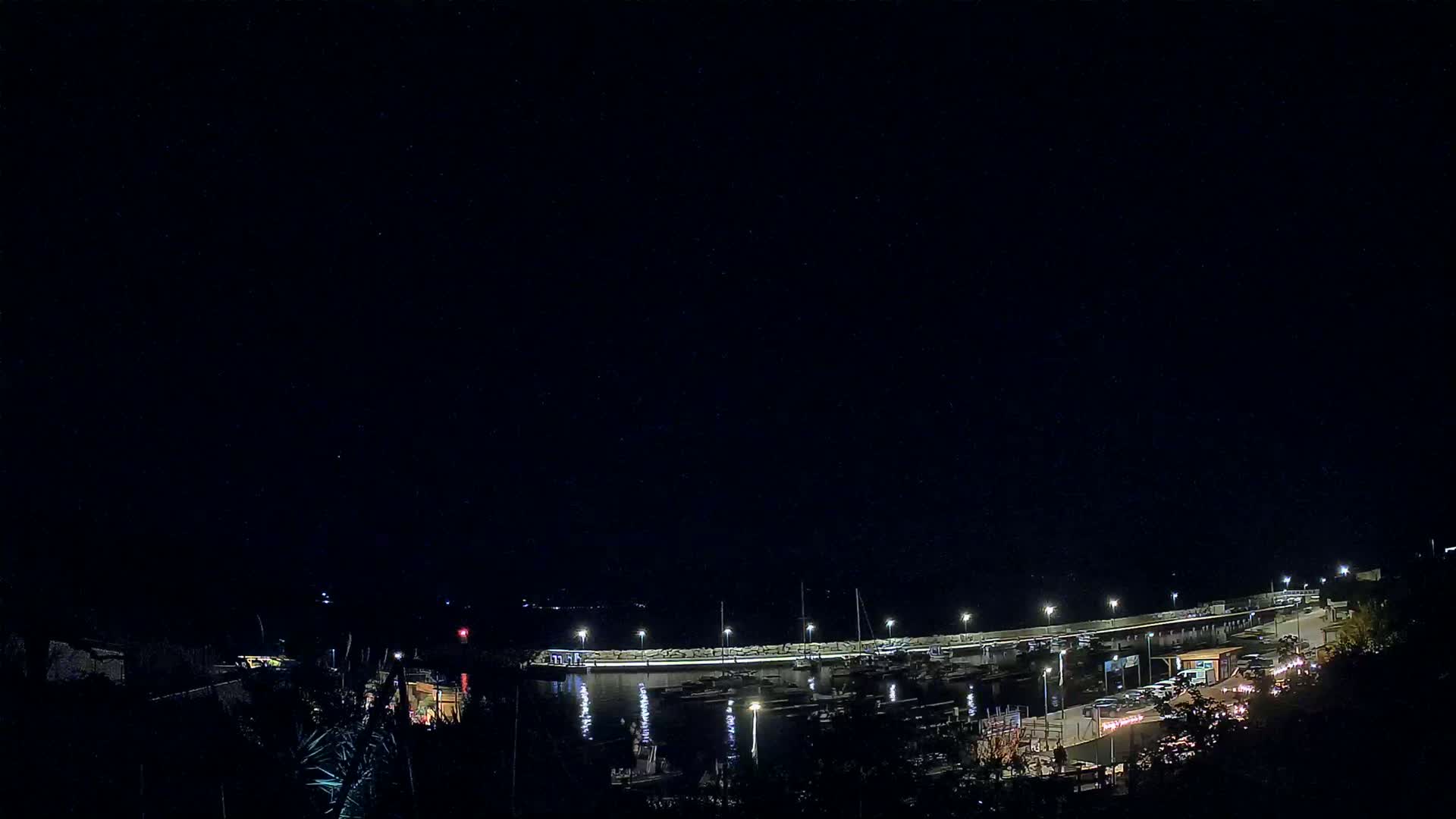 A nighttime view of a harbor filled with boats, illuminated by lights along a curved wall, under a clear, dark sky.