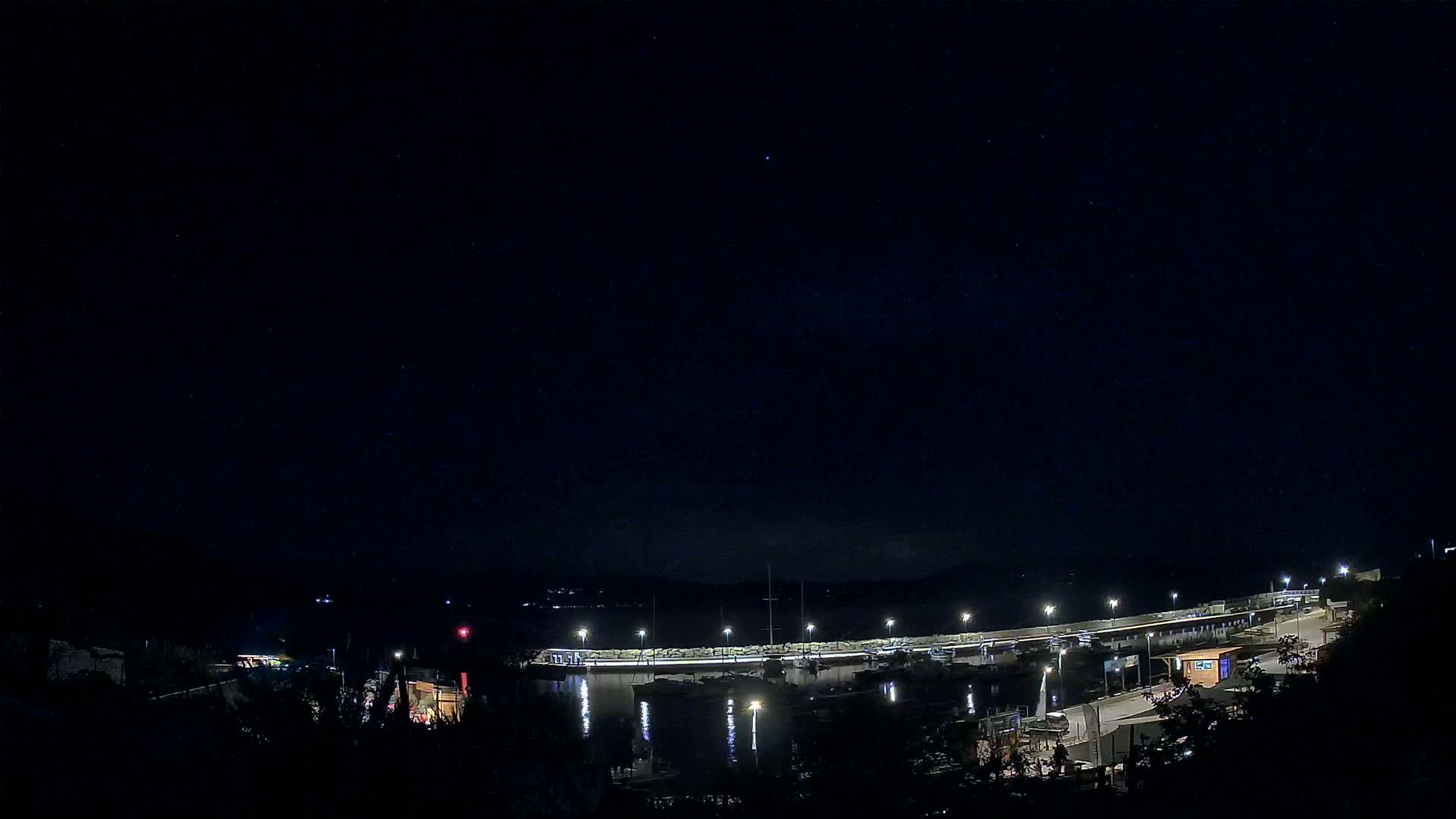 A nighttime view of a harbor filled with boats, illuminated by lights under a clear, starry sky.