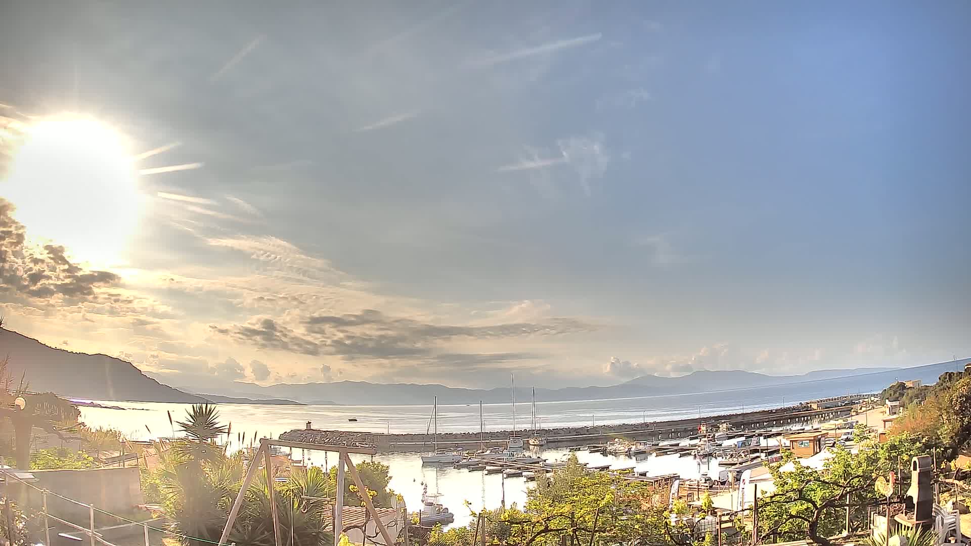 A partly cloudy sunny day overlooks a calm harbor filled with boats, with mountains visible in the distance.
