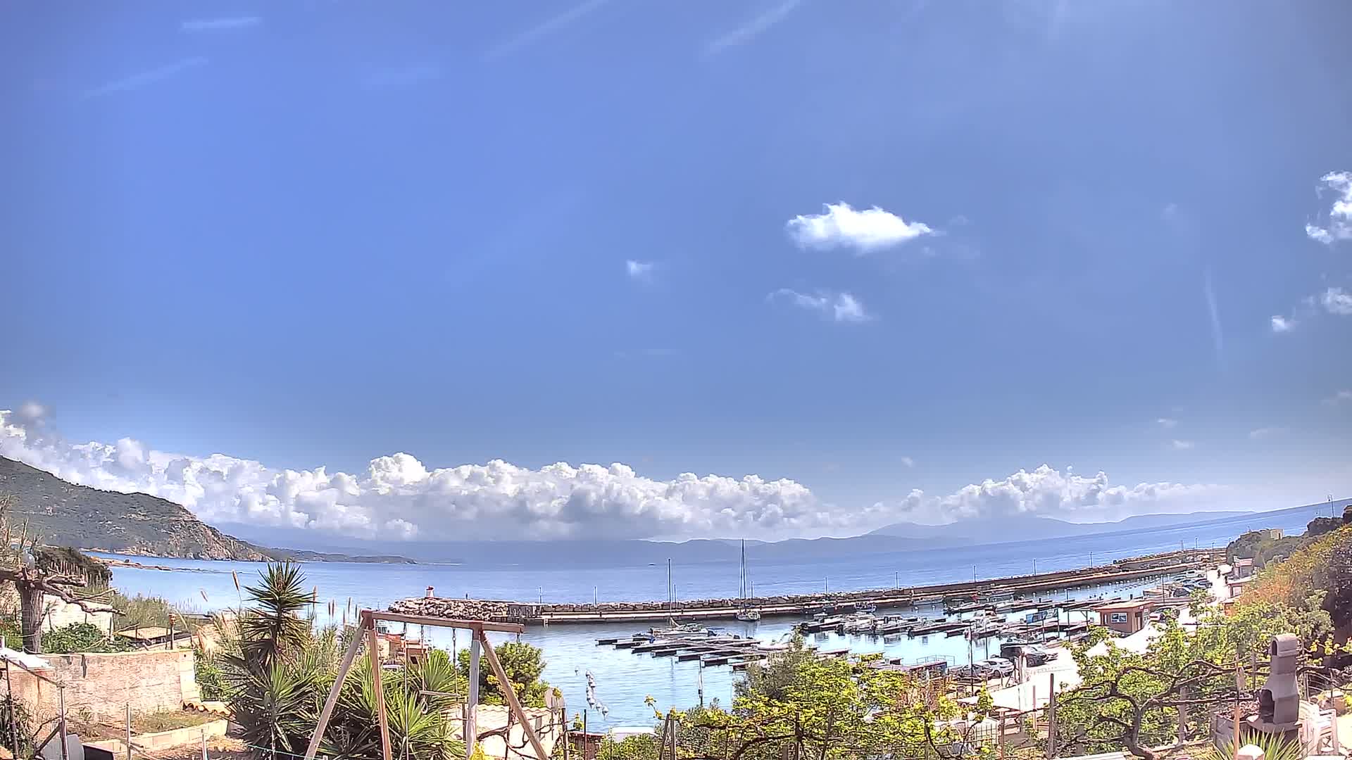 A small harbor filled with boats sits below a partly cloudy blue sky, with a calm sea extending to distant hills.