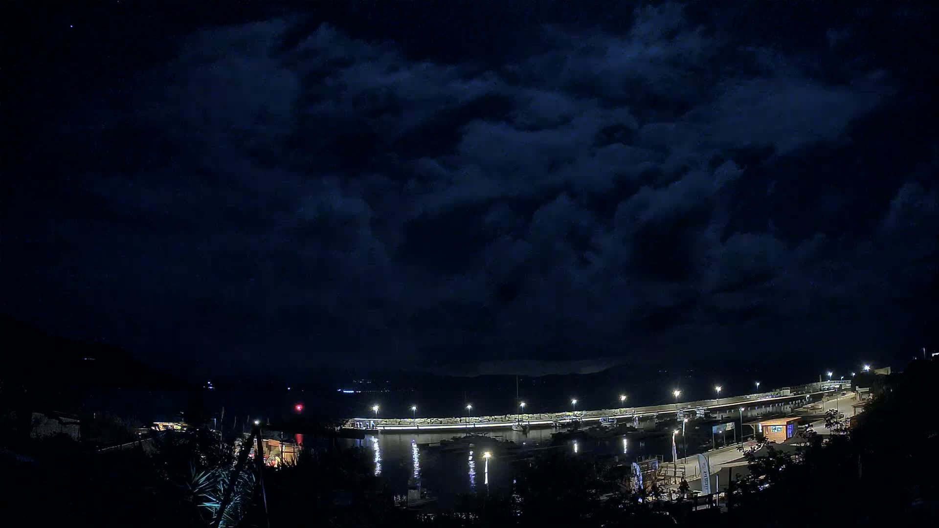 A nighttime view of a harbor with boats docked, illuminated by lights under a dark, cloudy sky.
