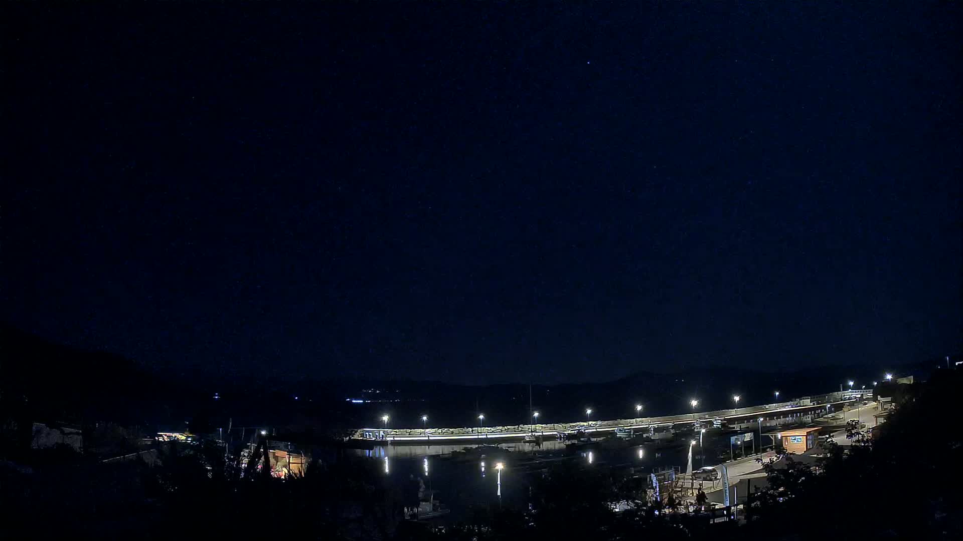A nighttime view of a harbor with boats docked, illuminated by lights against a dark, clear sky.