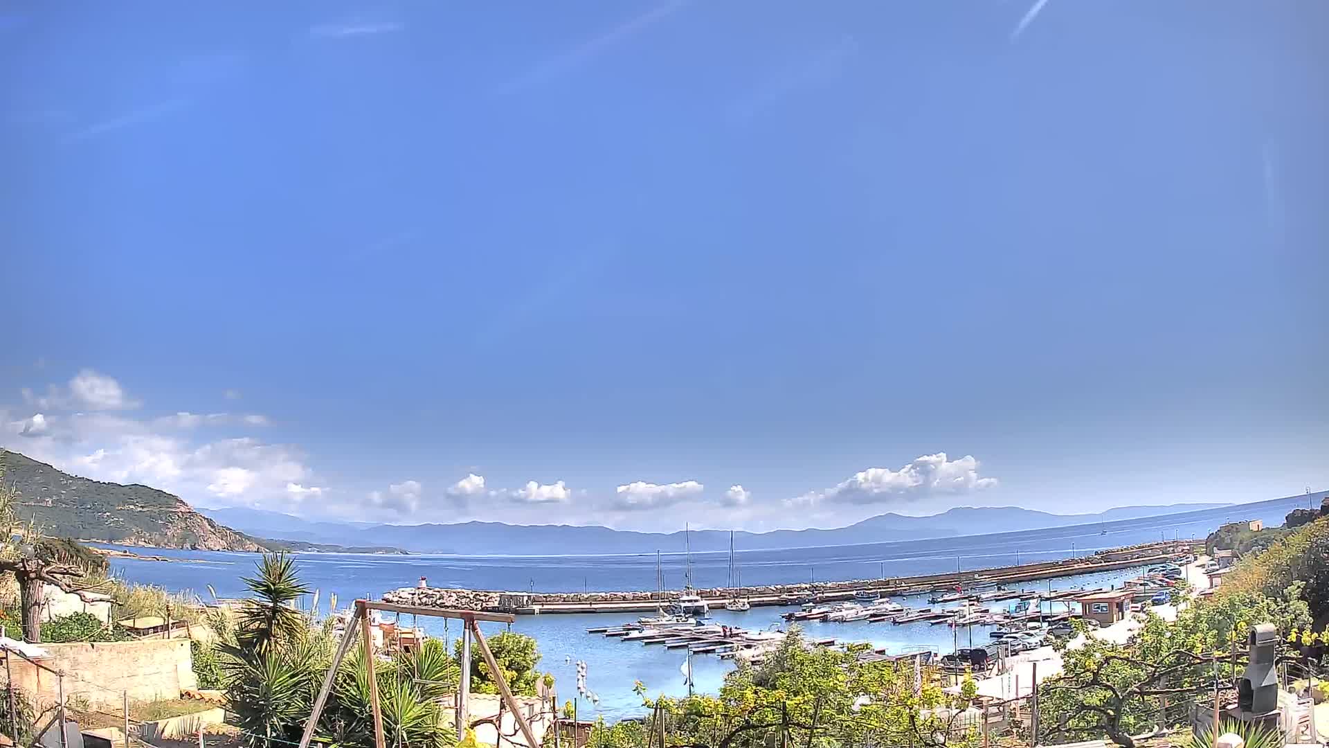 A mostly sunny day reveals a small harbor filled with boats, nestled between a hillside and a calm sea with distant mountains under a clear blue sky.