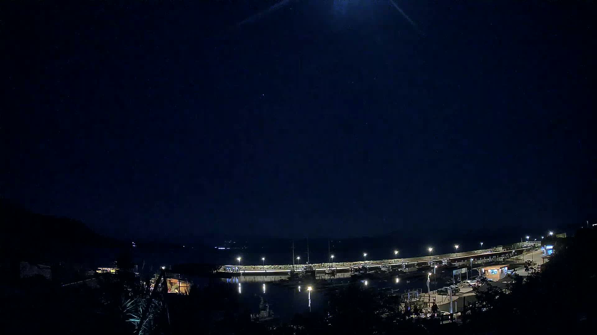 A moonlit night scene shows a harbor filled with boats, illuminated by lights along a seawall, with dark hillsides visible in the foreground and background.