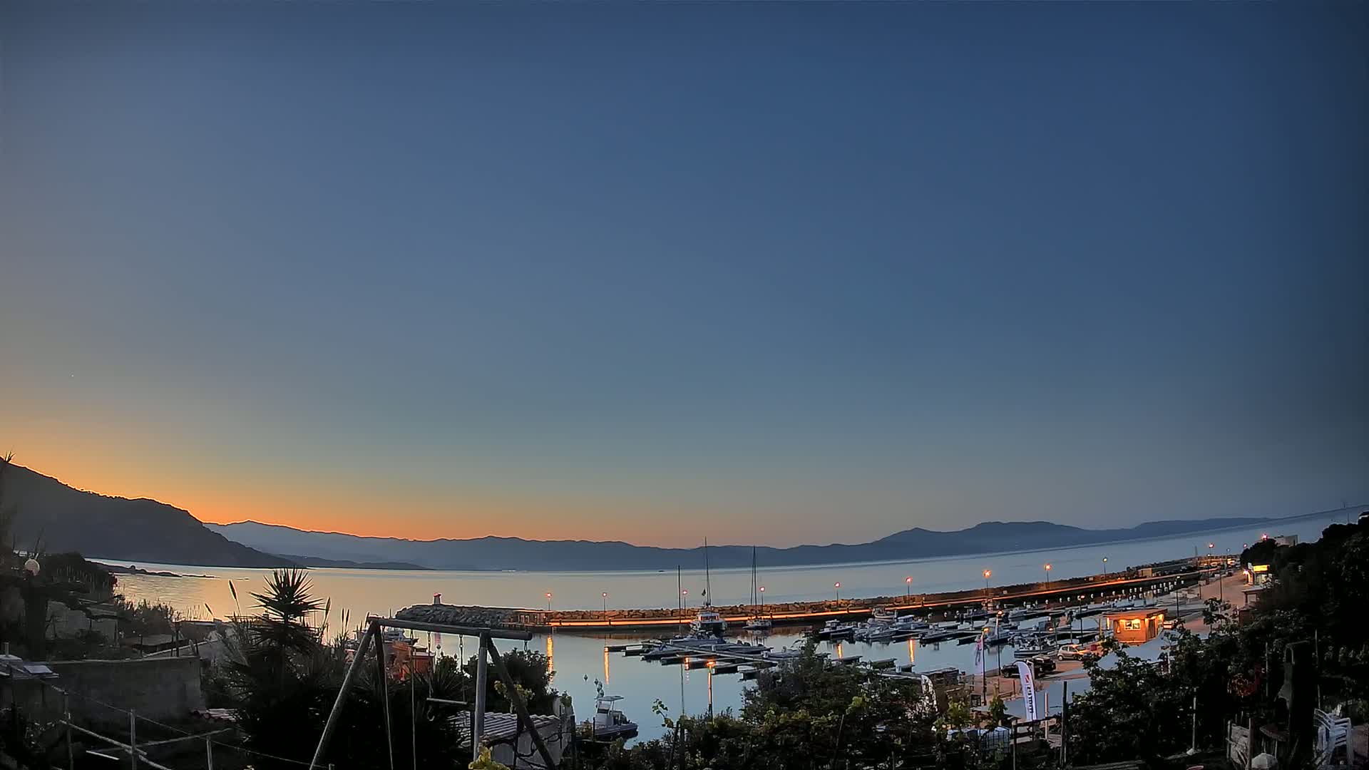 A tranquil harbor scene at sunset, featuring numerous boats moored at a pier, with calm waters reflecting the orange and blue hues of the twilight sky against a backdrop of silhouetted mountains.