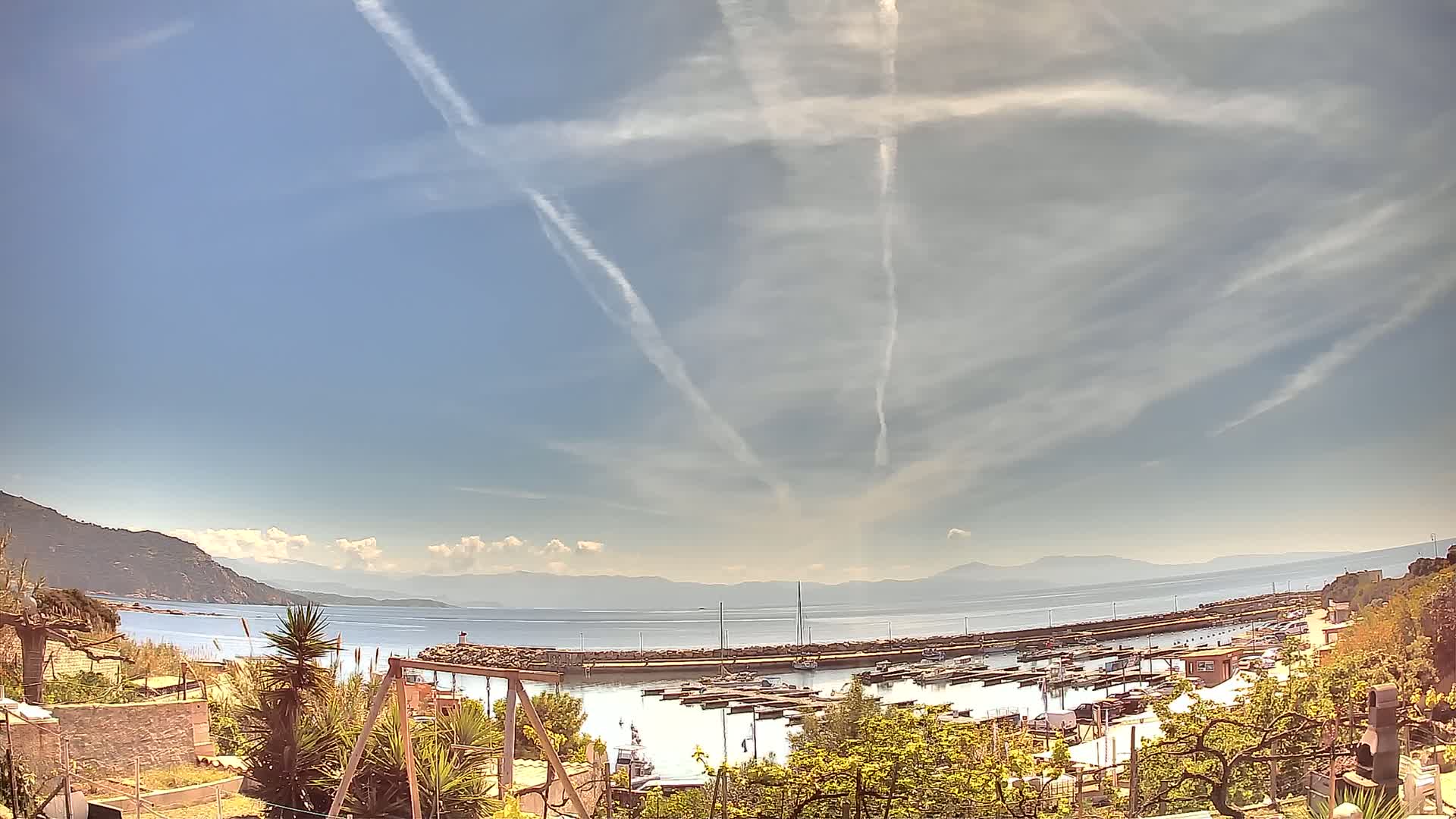 A marina filled with boats is visible from a hillside overlooking a calm sea under a mostly clear sky with contrails.