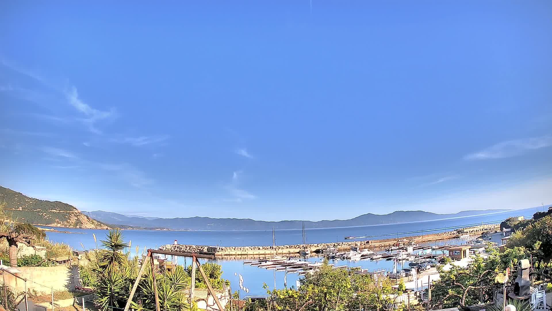 A small harbor filled with boats is nestled between a rocky hillside and calm, blue water under a mostly clear sky.