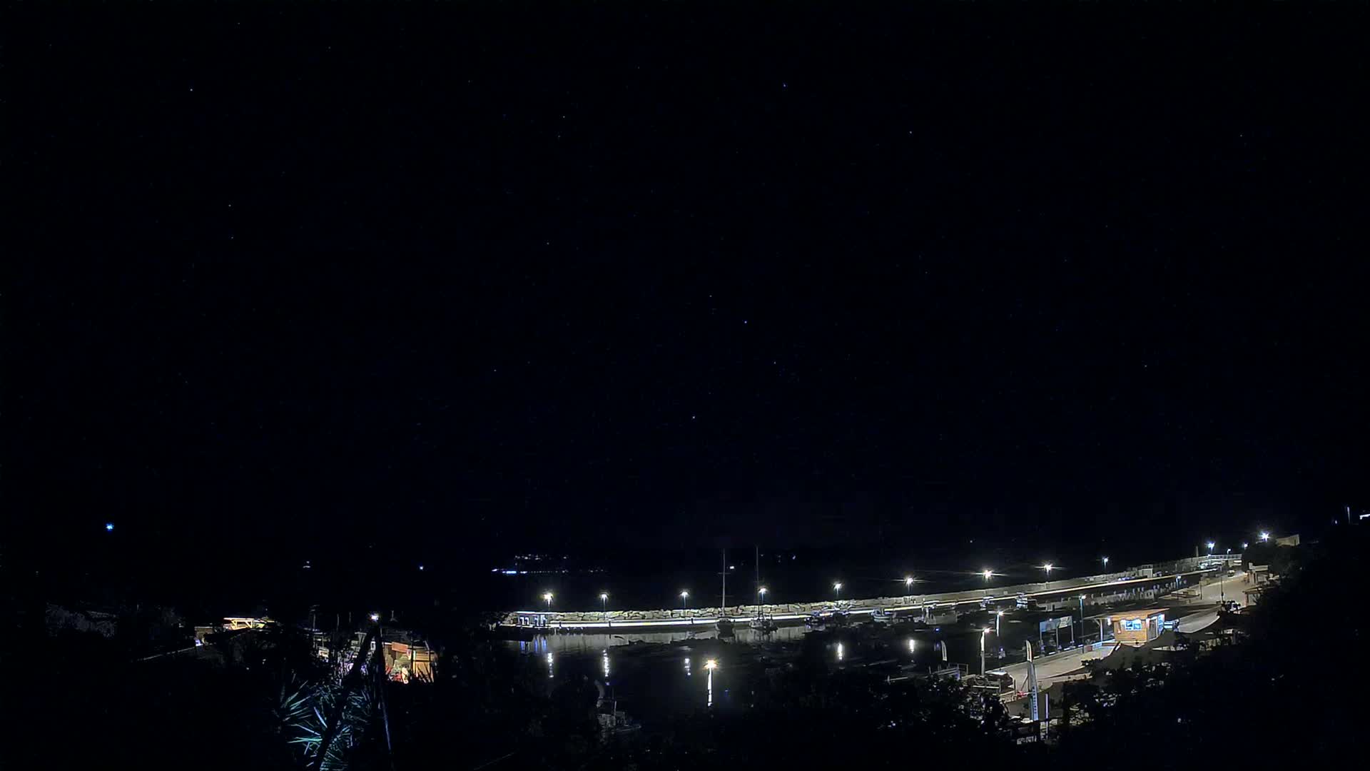 A nighttime view of a harbor filled with boats, lit by lamps along a breakwater under a clear, starry sky.