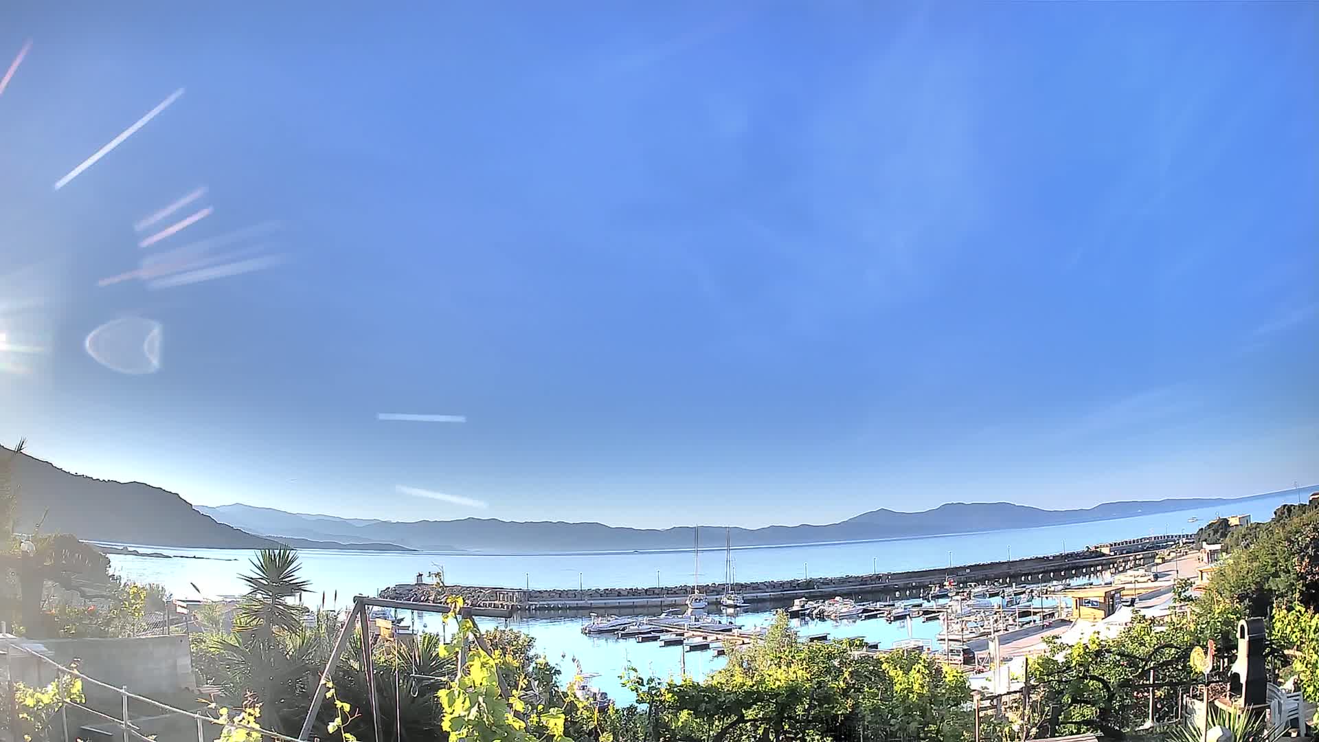 A sunny day reveals a harbor filled with boats, nestled between a tranquil bay and distant mountains.