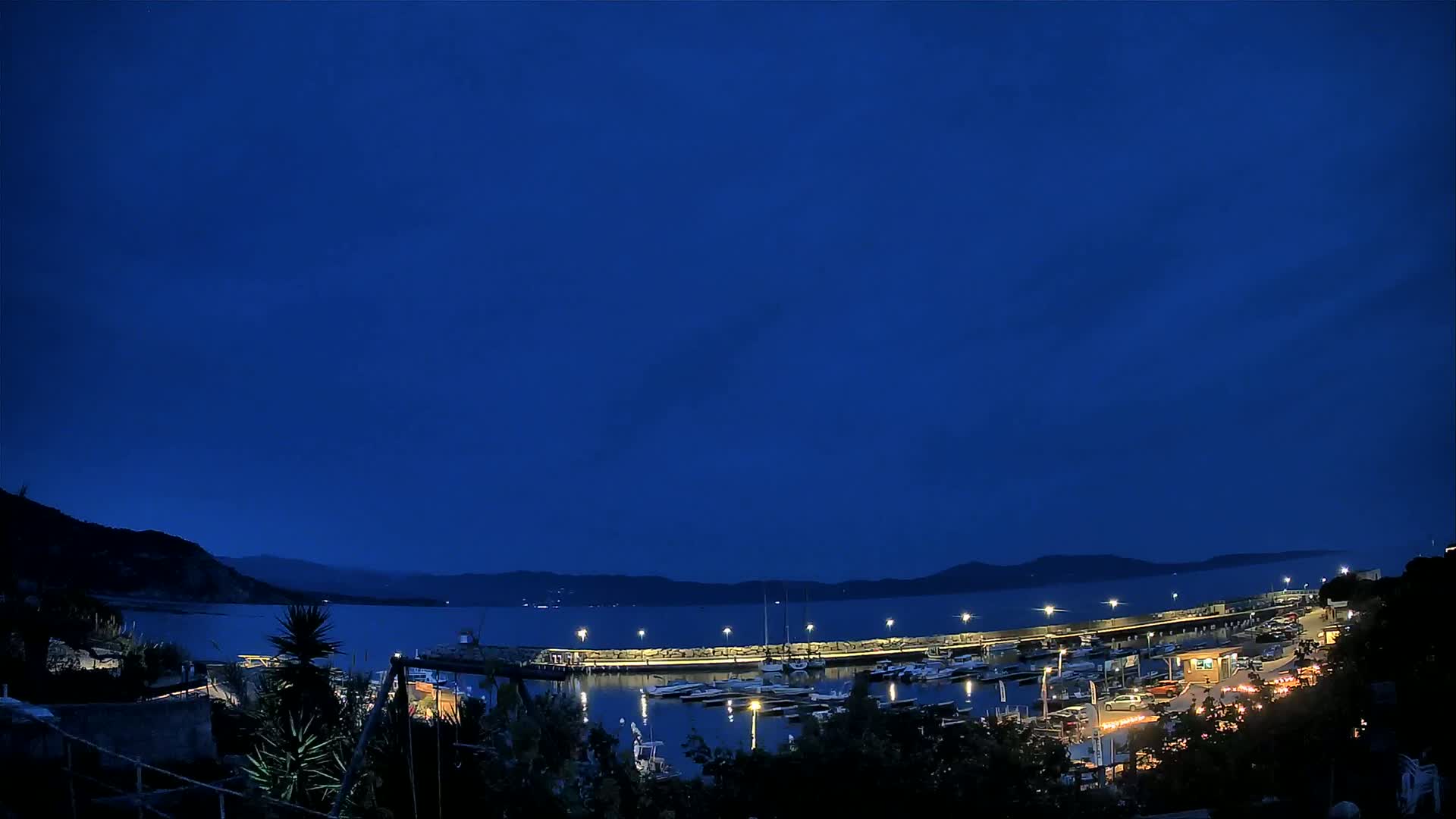 A nighttime view of a harbor filled with boats, illuminated by lights against a dark blue sky.