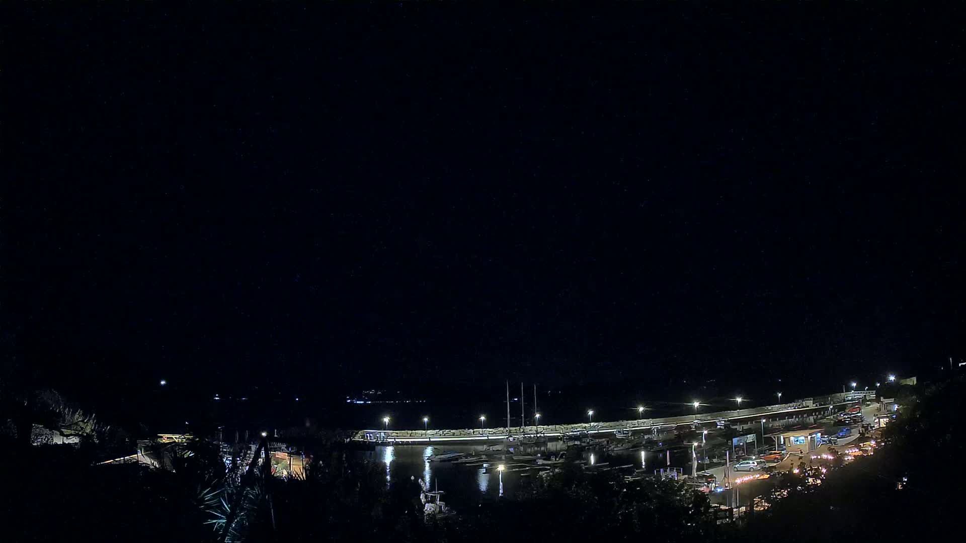 A nighttime view of a harbor filled with boats, illuminated by lights along a dock, under a clear, dark sky.