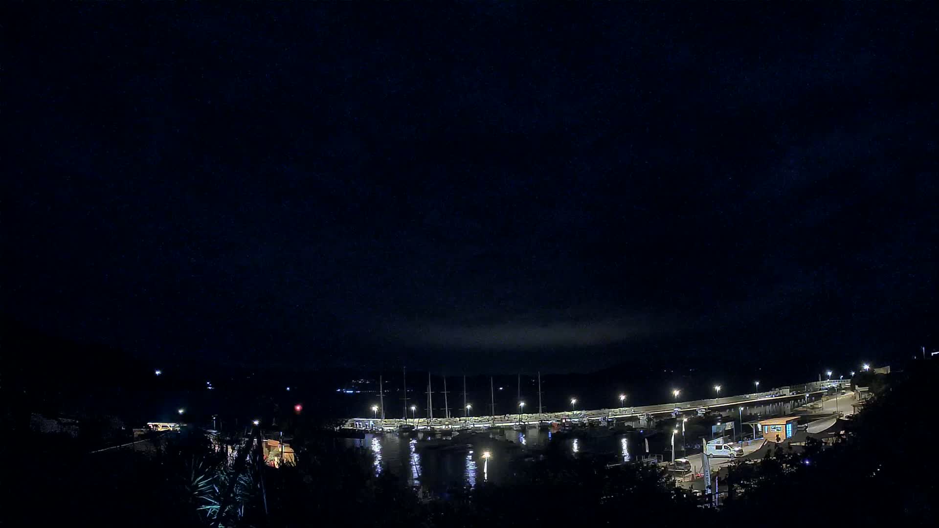A nighttime view of a marina with boats docked, illuminated by lights under a dark, mostly clear sky.