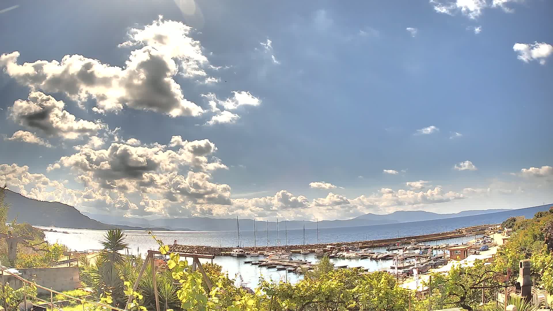 A partly sunny day shows a marina filled with boats, nestled between a hillside of vegetation and a calm bay backed by distant mountains.