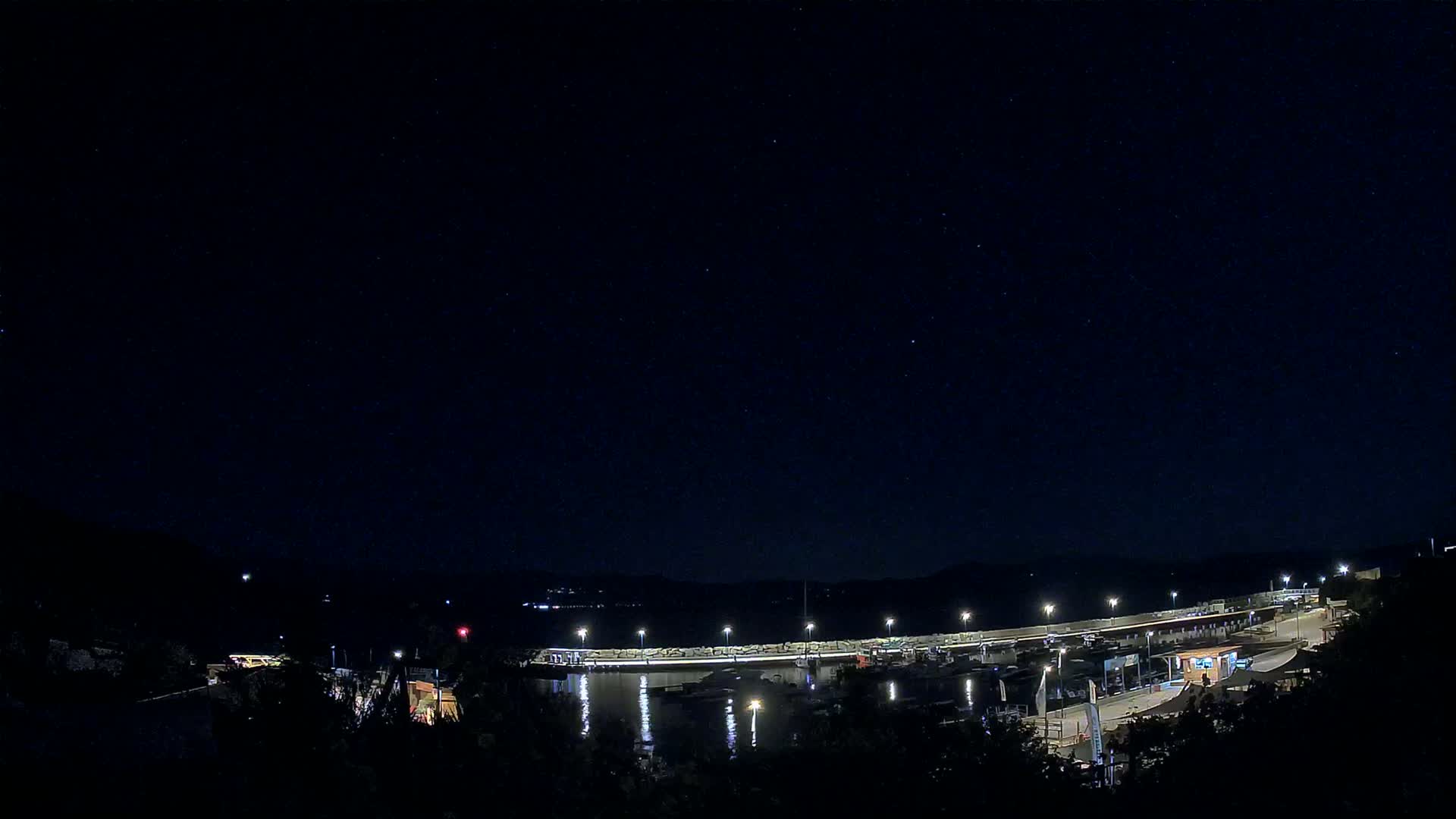 Under a clear, starry night sky, a marina with docked boats is illuminated, surrounded by dark hills.