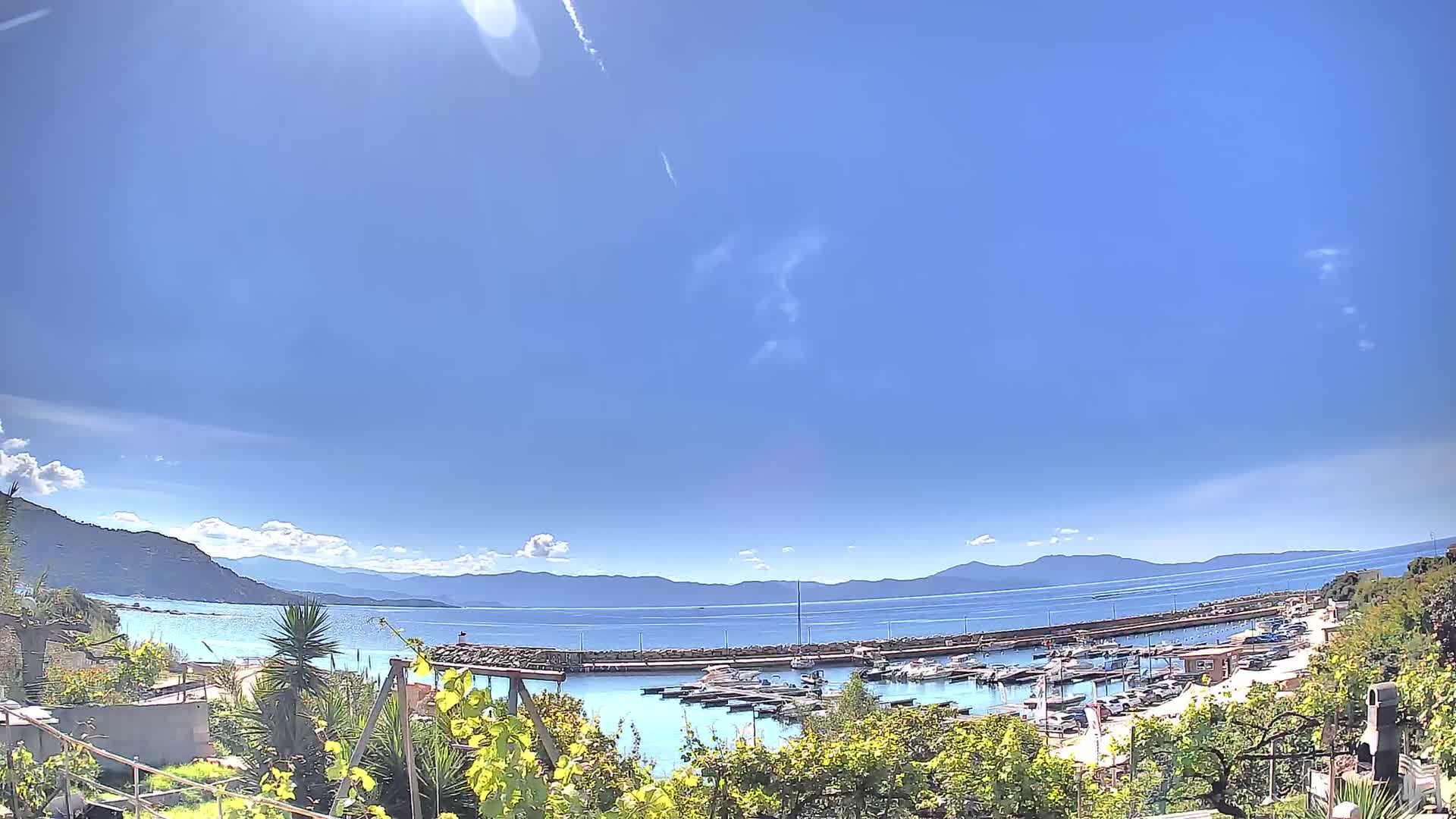 A sunny day reveals a harbor filled with boats, nestled between a verdant hillside and a calm, blue sea, backed by distant mountains under a mostly clear sky.