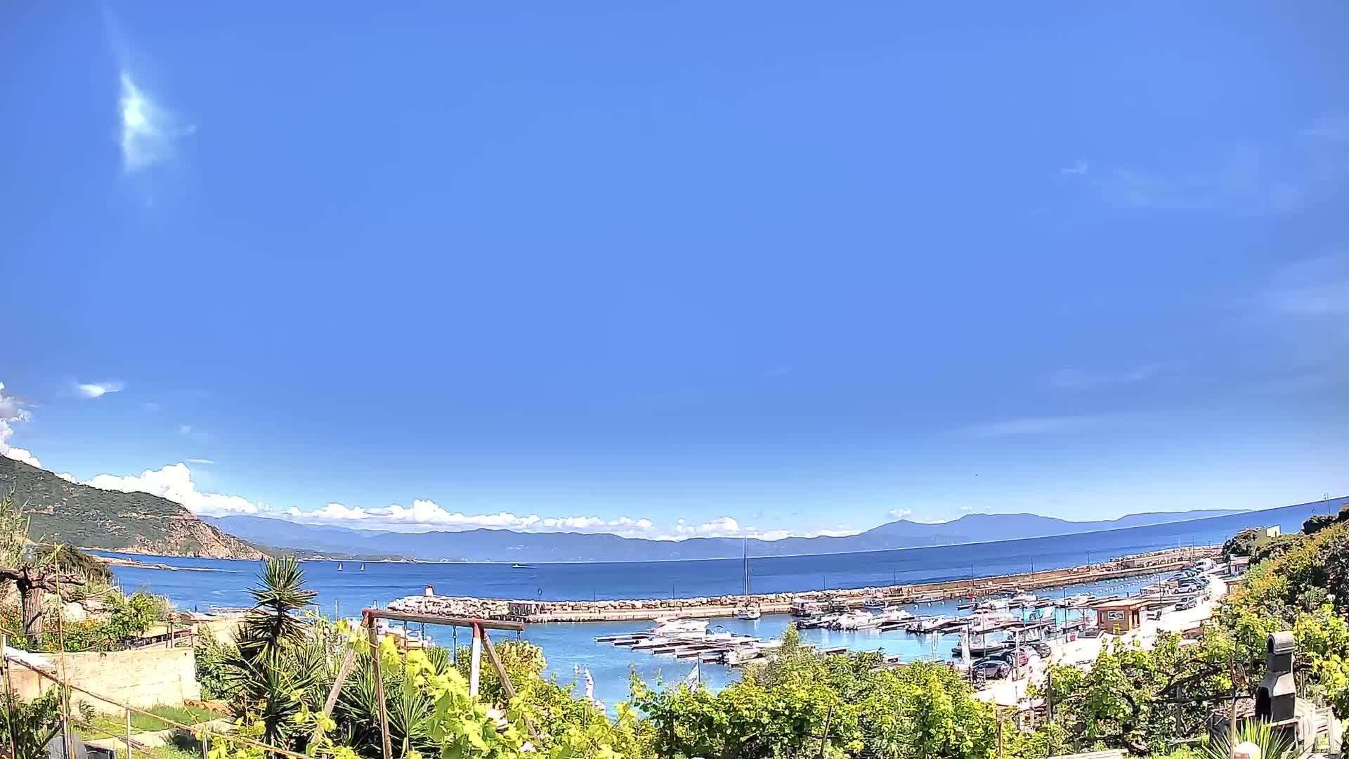 A marina filled with boats is nestled between a verdant hillside and a calm, blue sea under a mostly clear, sunny sky.