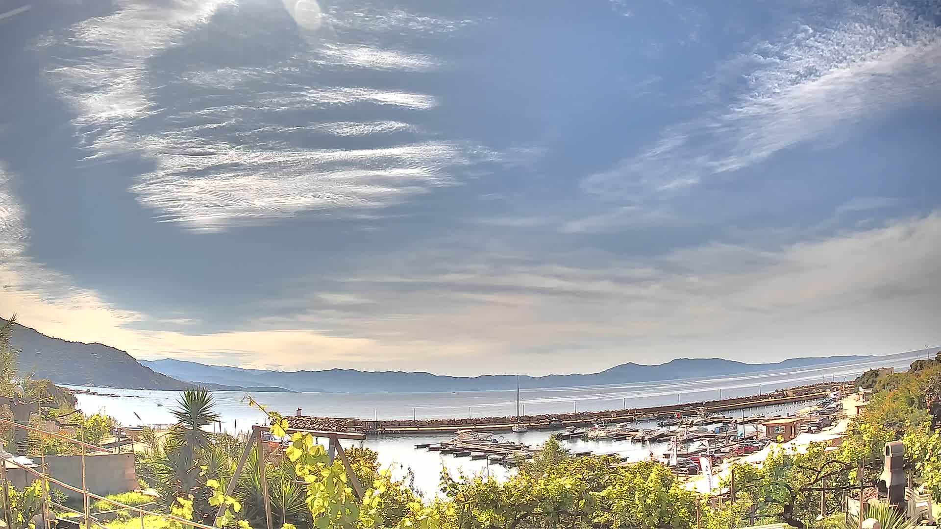 A partly sunny day reveals a marina filled with boats, situated between a verdant hillside and a calm body of water extending to distant mountains under a mostly clear sky.