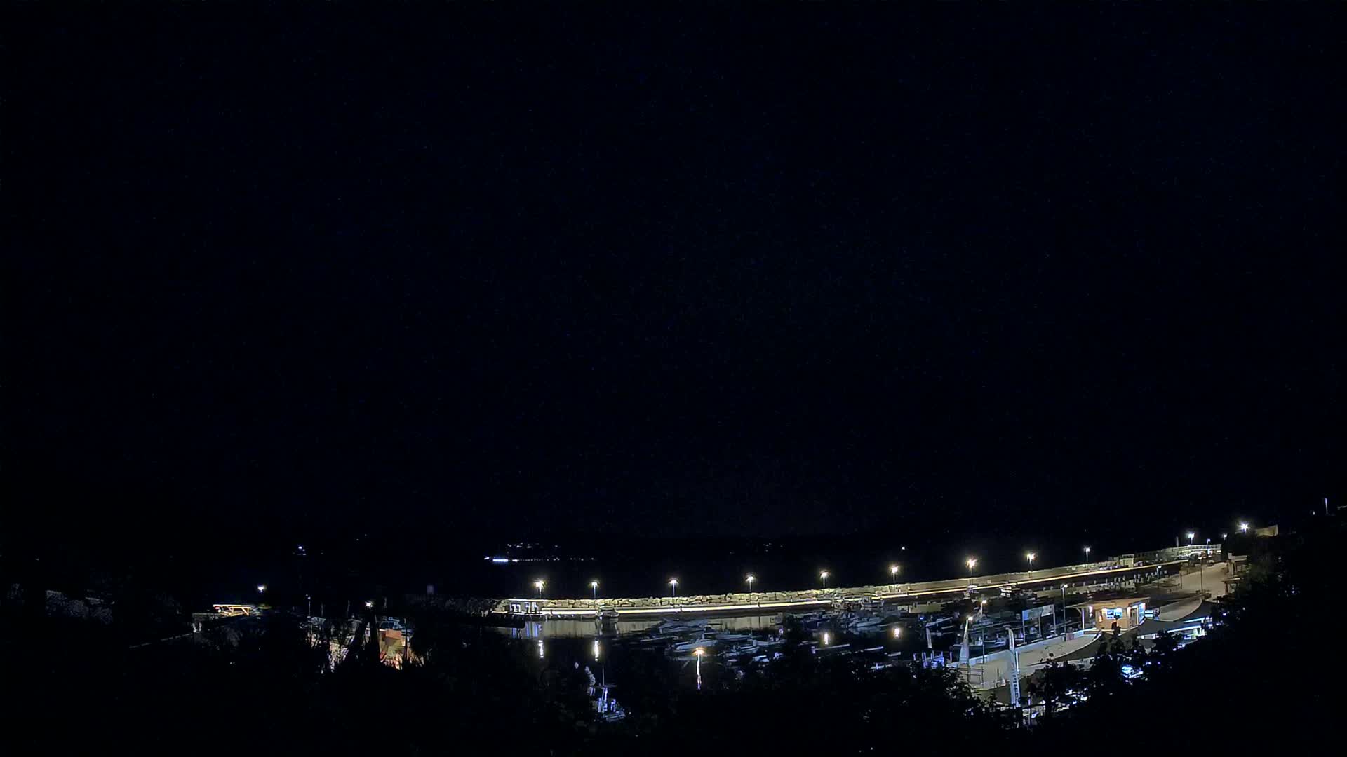 A nighttime view of a marina with many boats docked, illuminated by lights under a clear, dark sky.