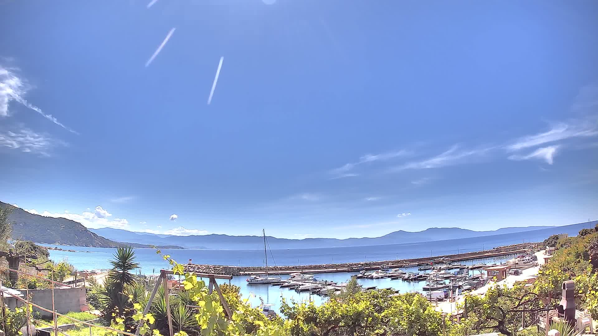 A sunny day reveals a calm harbor filled with boats, nestled between a coastal town and a mountainous backdrop.