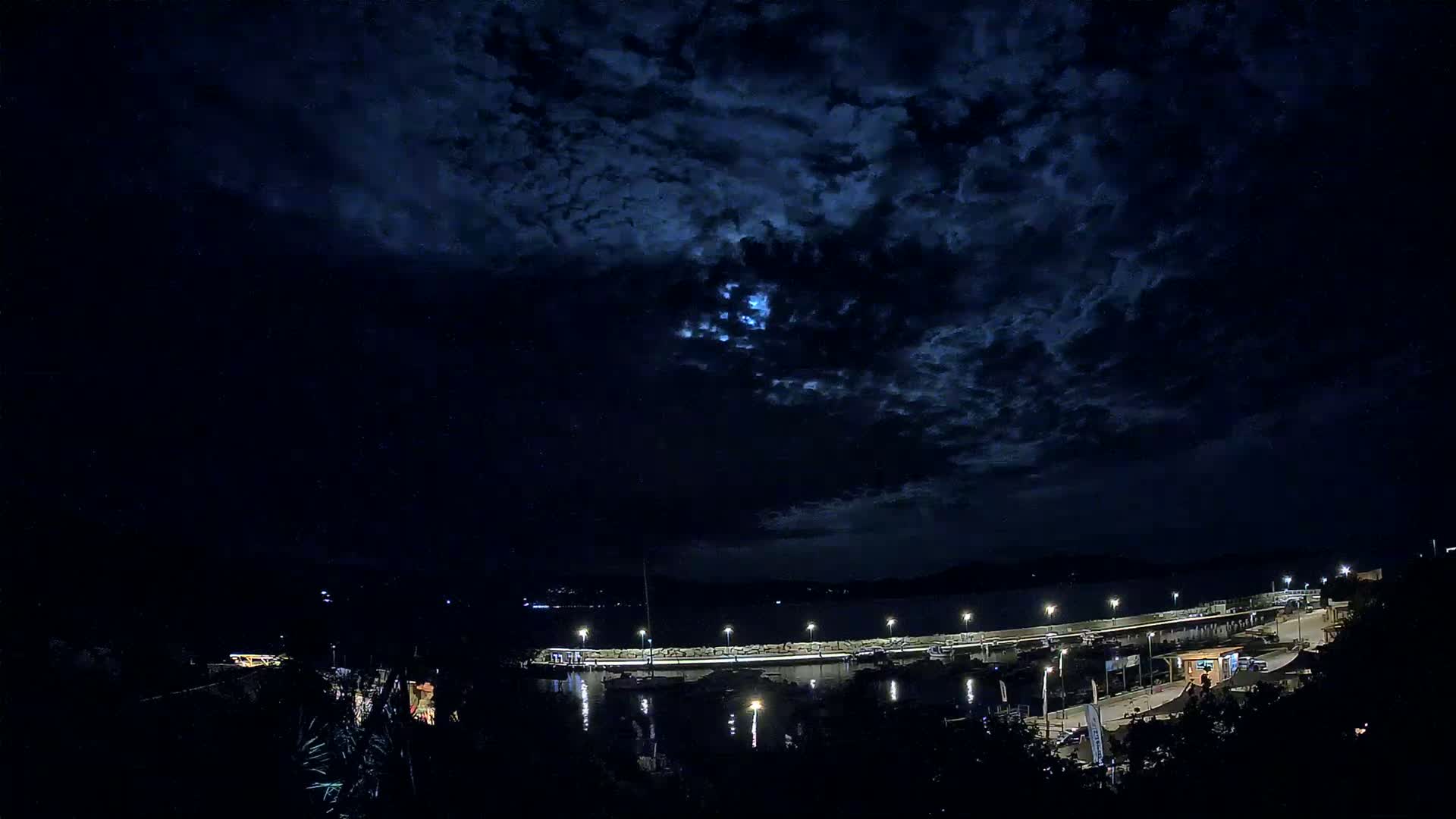A moonlit nighttime scene shows a harbor with boats and lights under a partly cloudy sky.