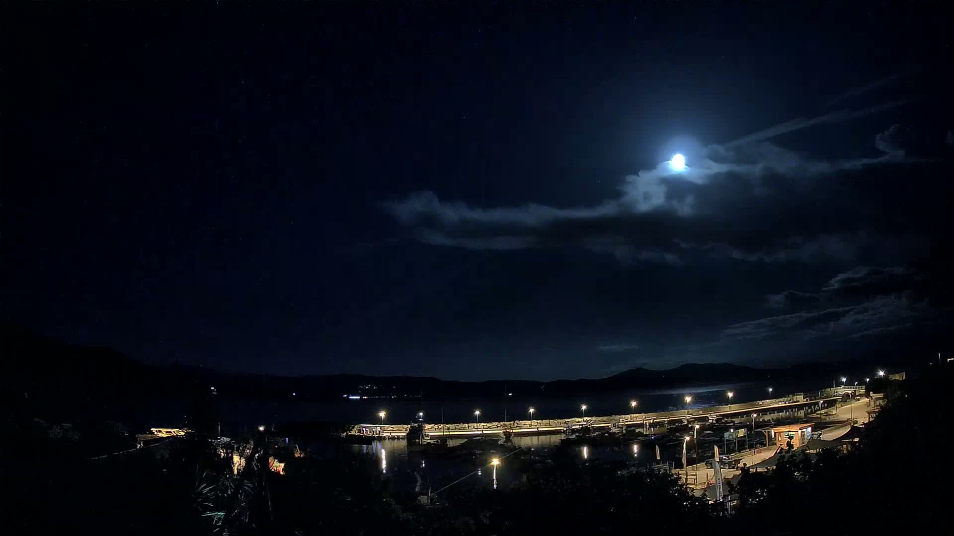 A brightly lit harbor with boats is visible under a mostly cloudy night sky illuminated by a bright moon.