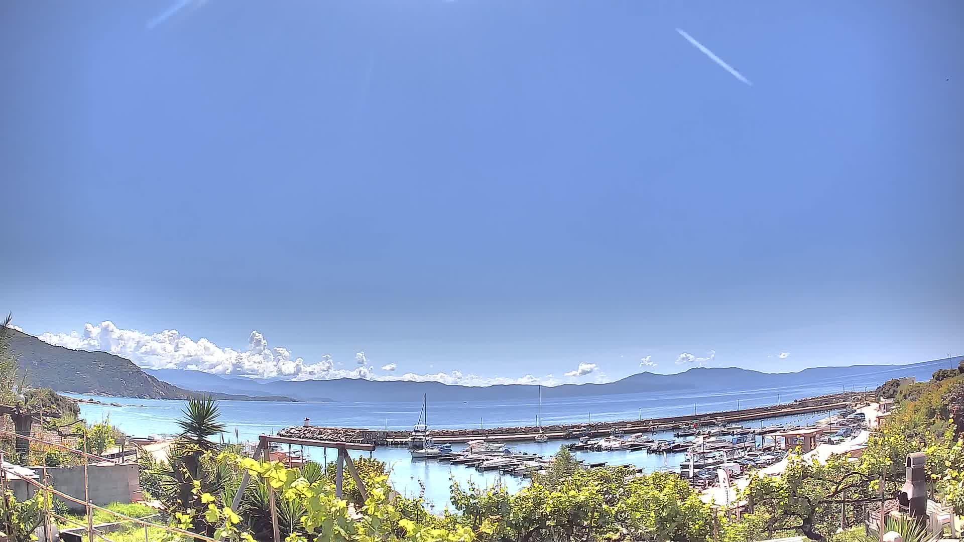 A marina filled with boats is nestled in a calm bay, under a mostly clear blue sky with a few fluffy clouds, backed by distant mountains.